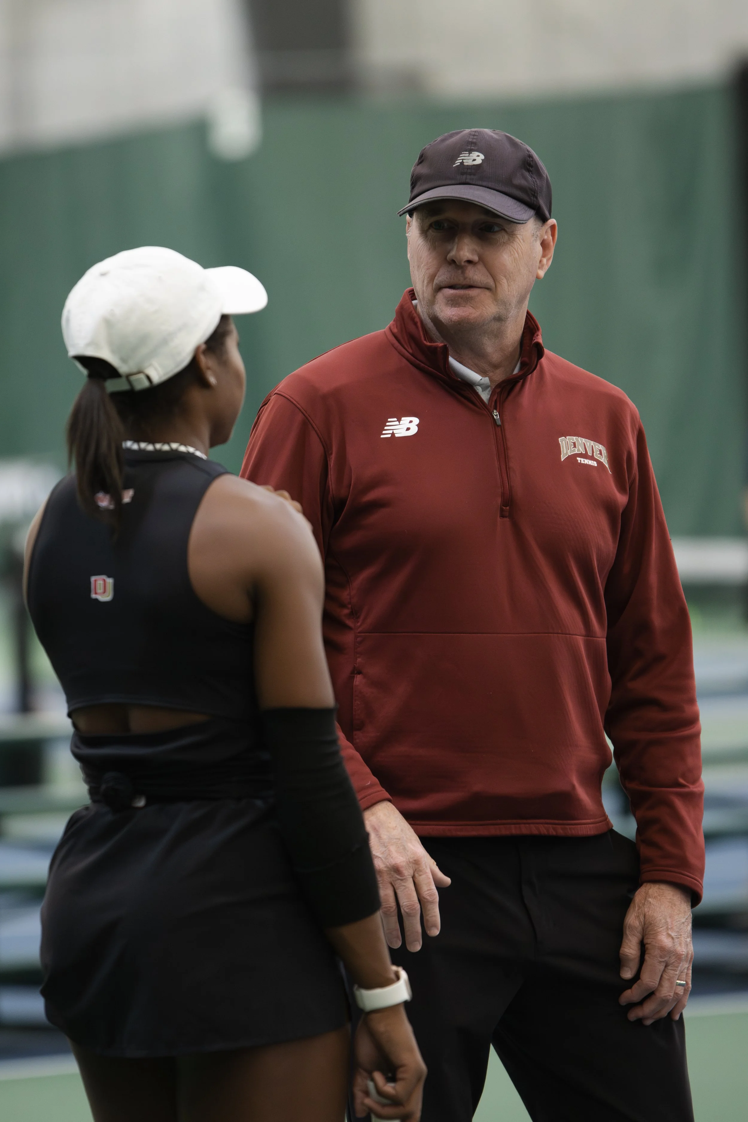 A male tennis coach and a female tennis player having a conversation on an indoor tennis court.