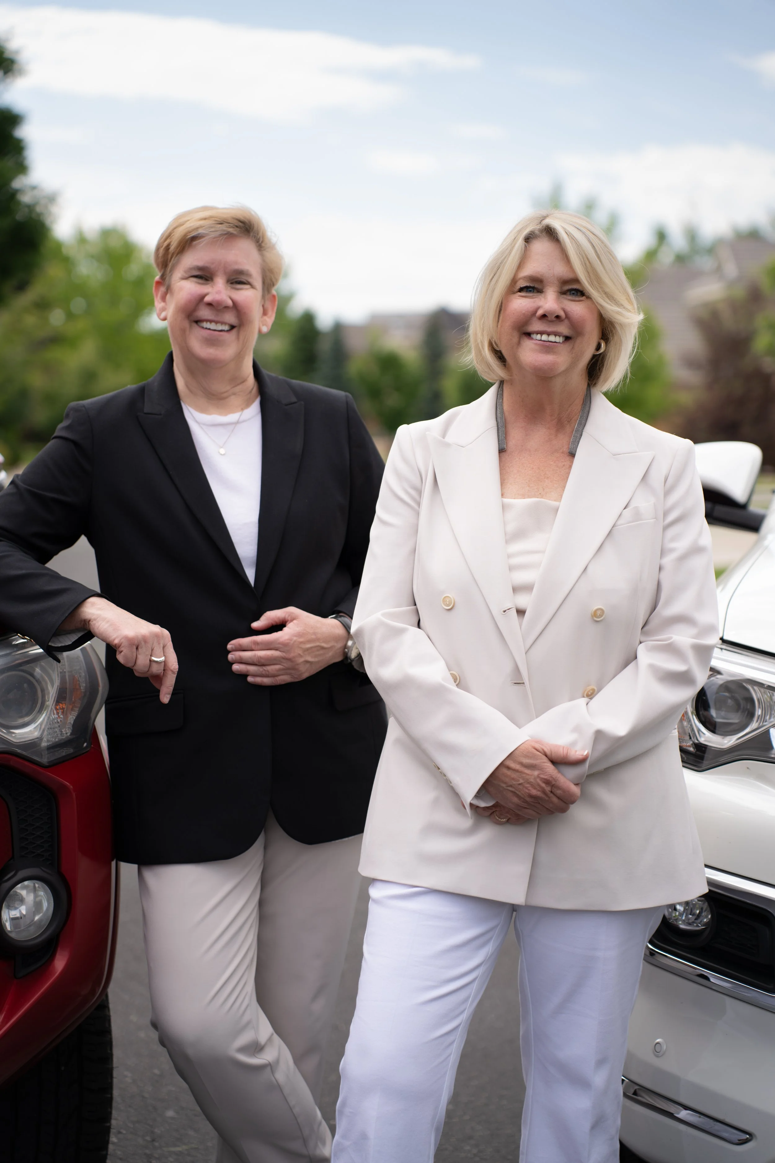 Two women standing outdoors next to cars, smiling at the camera. One woman is wearing a black blazer and white pants, the other is dressed in a white blazer and white pants.