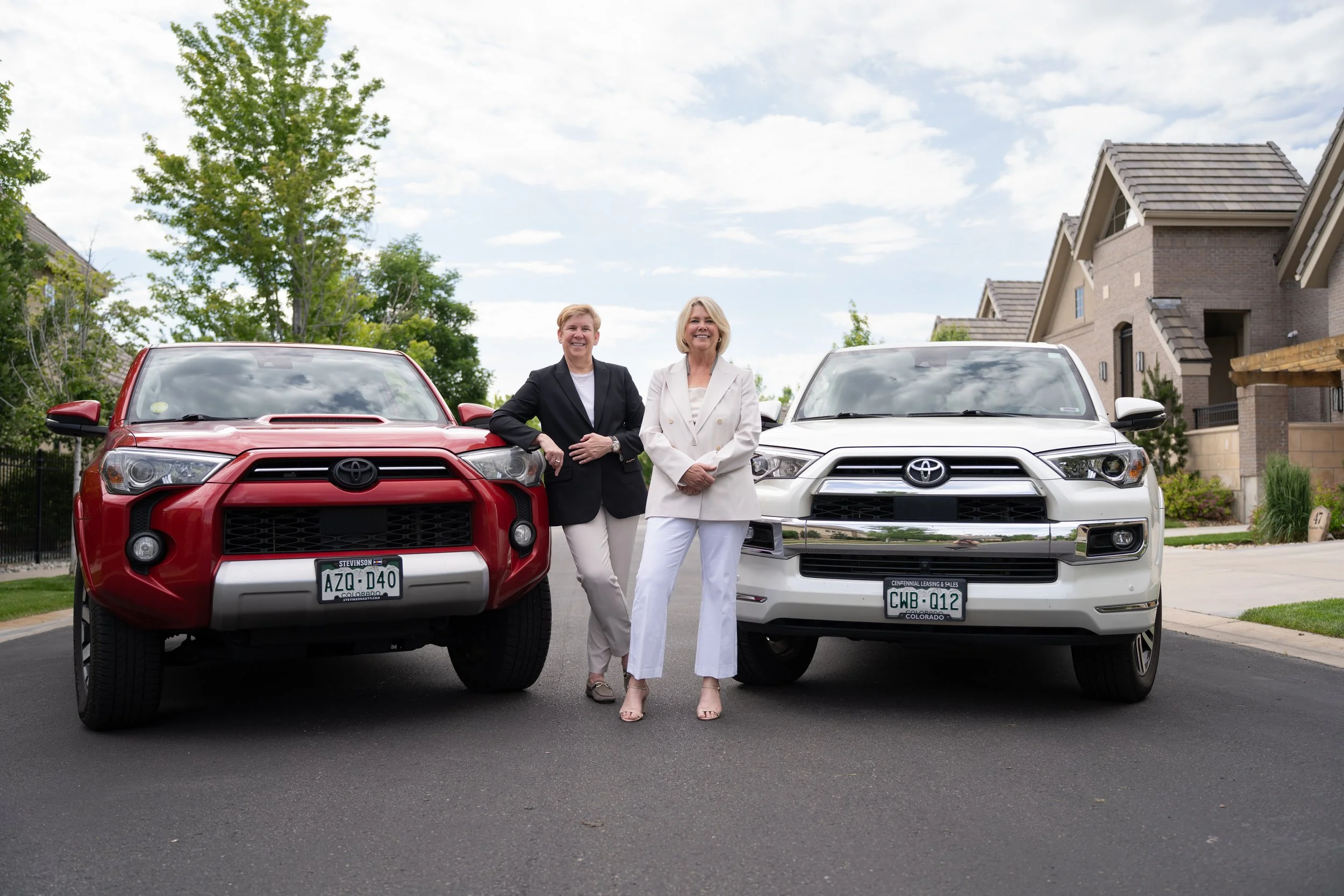 Two women standing between a red SUV and a white SUV on a residential street, smiling and posing for the camera.