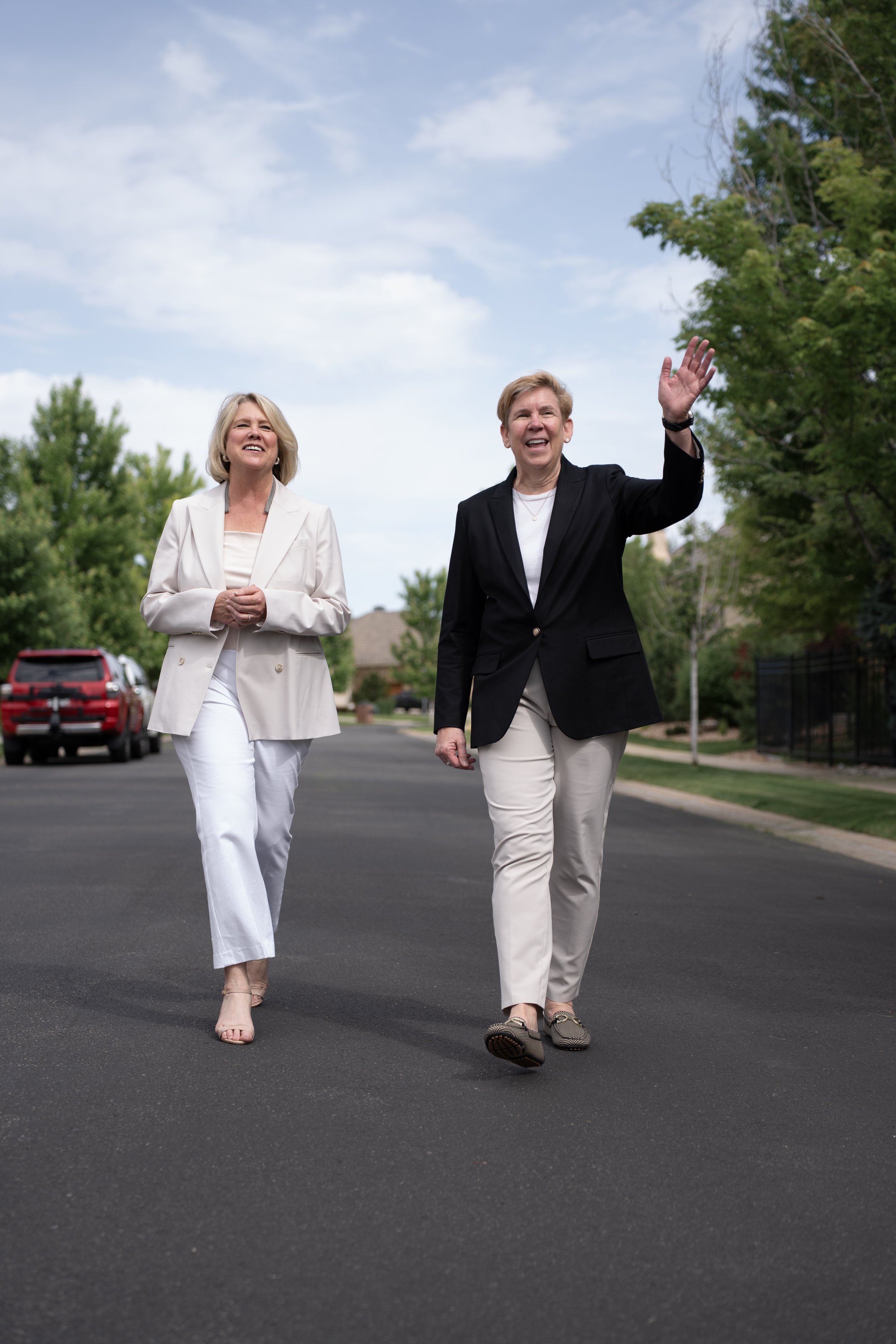 Two women walking and smiling on a suburban street, one waving, surrounded by green trees and parked cars.