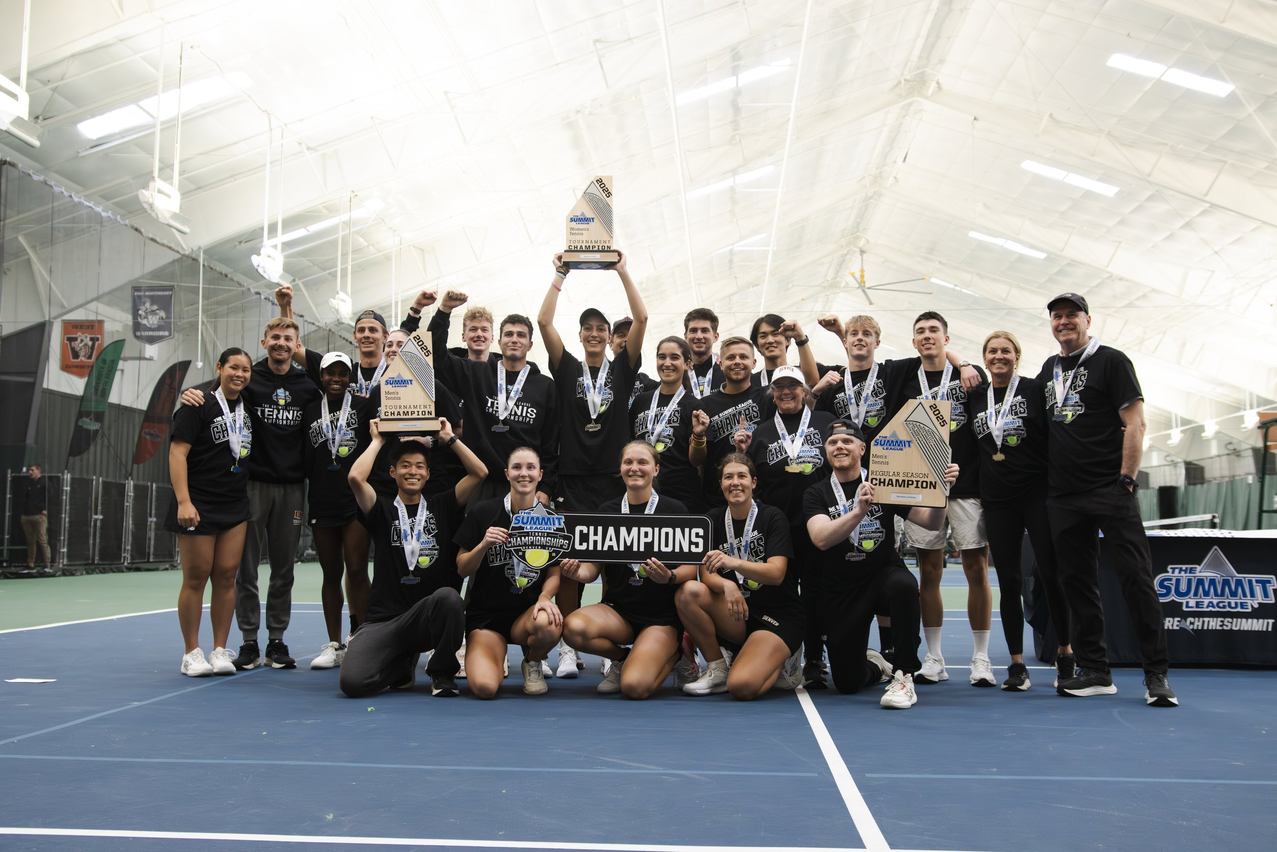 A tennis team celebrating after winning the championship on an indoor court, holding trophies and a 'Champions' sign, all wearing medals and black t-shirts with championship logos.