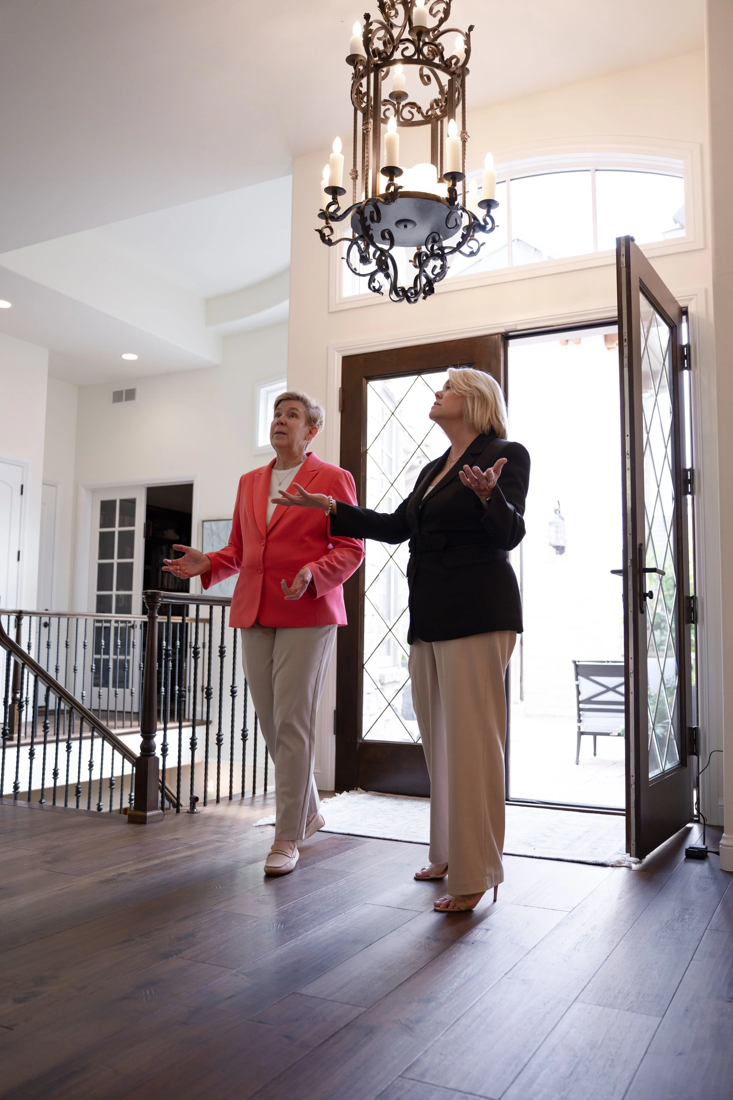 Two women standing inside a home near an open front door, engaged in a conversation. One woman is wearing a black blazer and beige pants, gesturing with her hands, while the other woman is wearing a red blazer and beige pants, listening and looking u