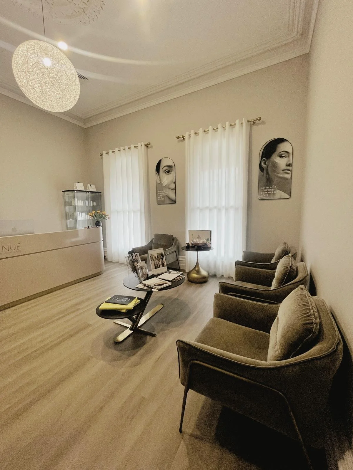 A waiting room with four beige armchairs, a small black coffee table with magazines and a book, two windows with white curtains, black and white portraits on the wall, a reception desk, and a glass display case.