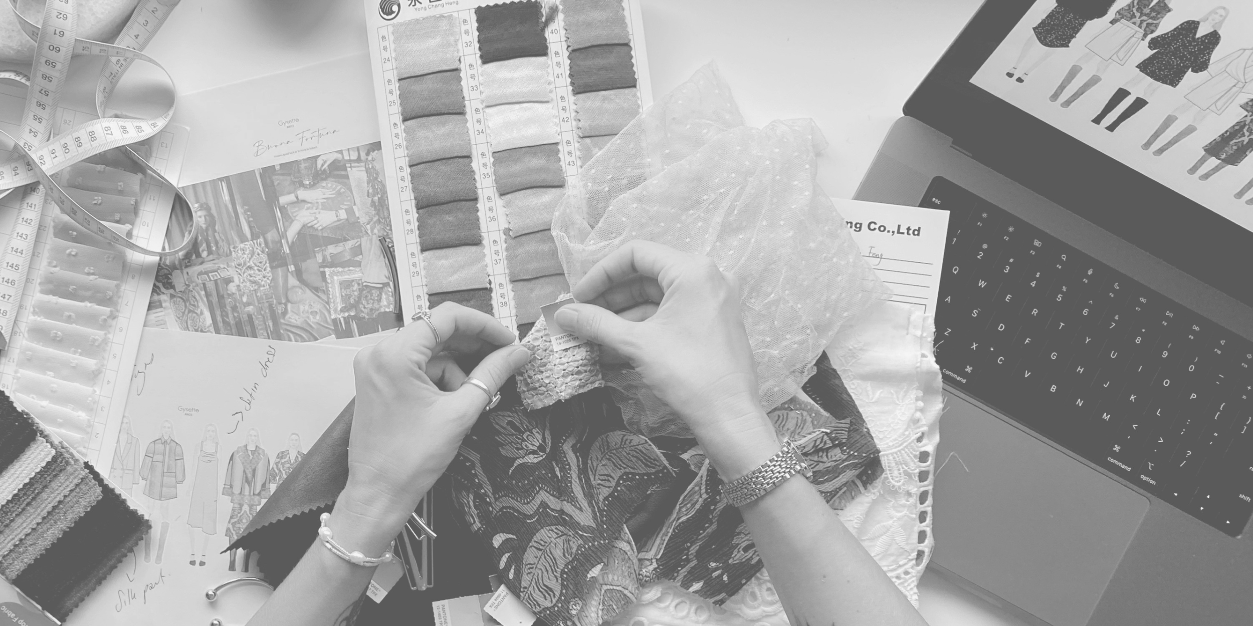 A person working on fabric samples and swatches at a desk with a laptop, design sketches, measuring tape, and fabric options surrounding them.