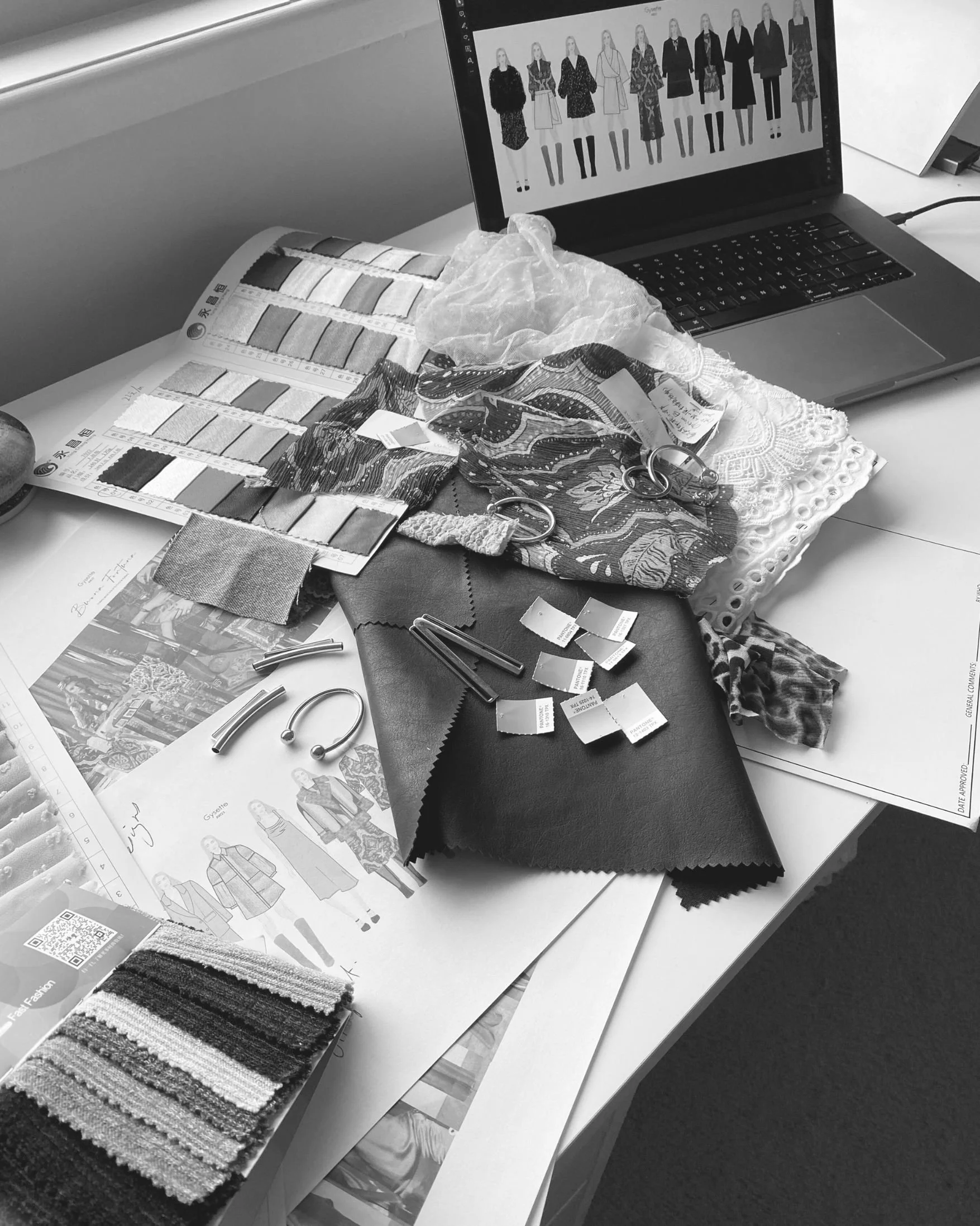 A workspace with fabric swatches, color samples, sewing patterns, scissors, a laptop displaying fashion sketches, and dress fabric samples.