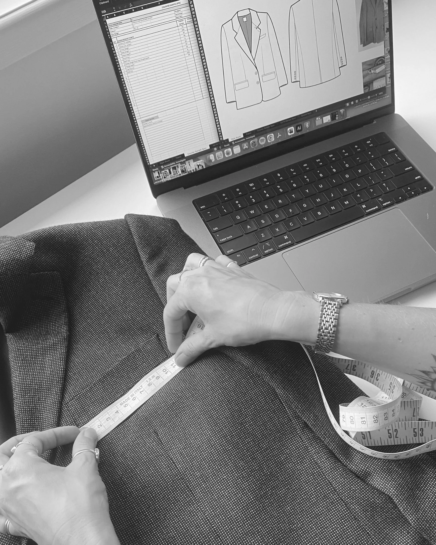 A person is measuring the waist of fabric with a tape measure while sitting at a desk with a laptop displaying fashion sketches of suits.