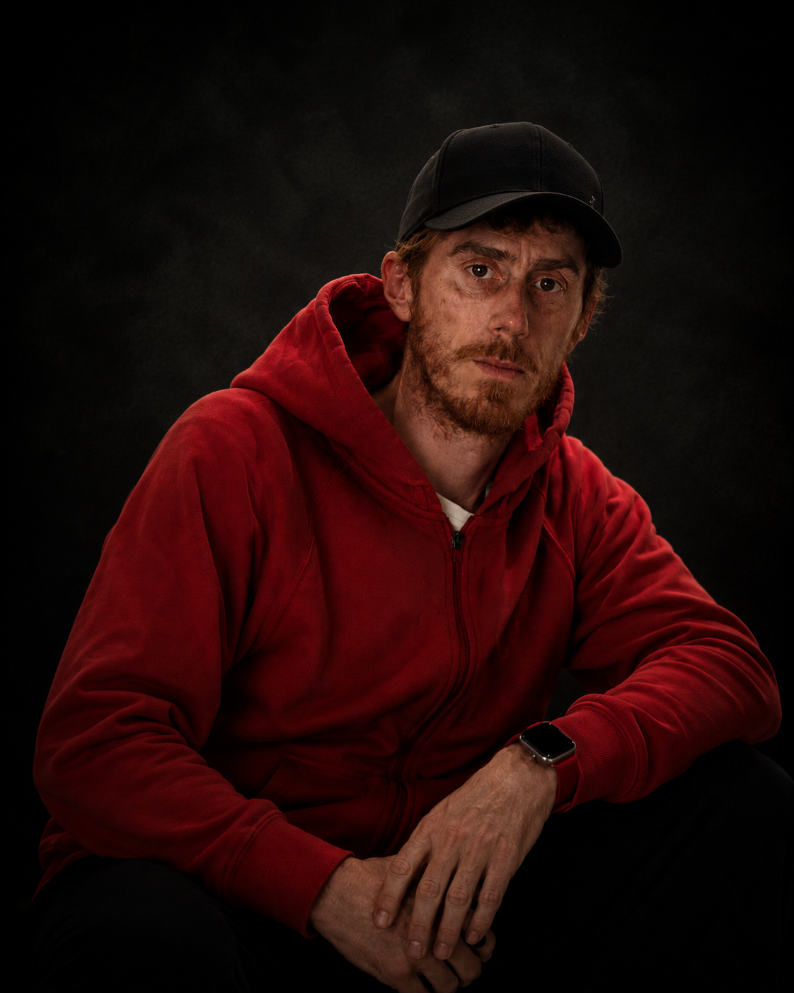 Ritratto di un uomo con cappellino nero, maglietta bianca e felpa rossa, seduto contro sfondo nero, con espressione seria e braccio appoggiato su un ginocchio, indossando un smartwatch.