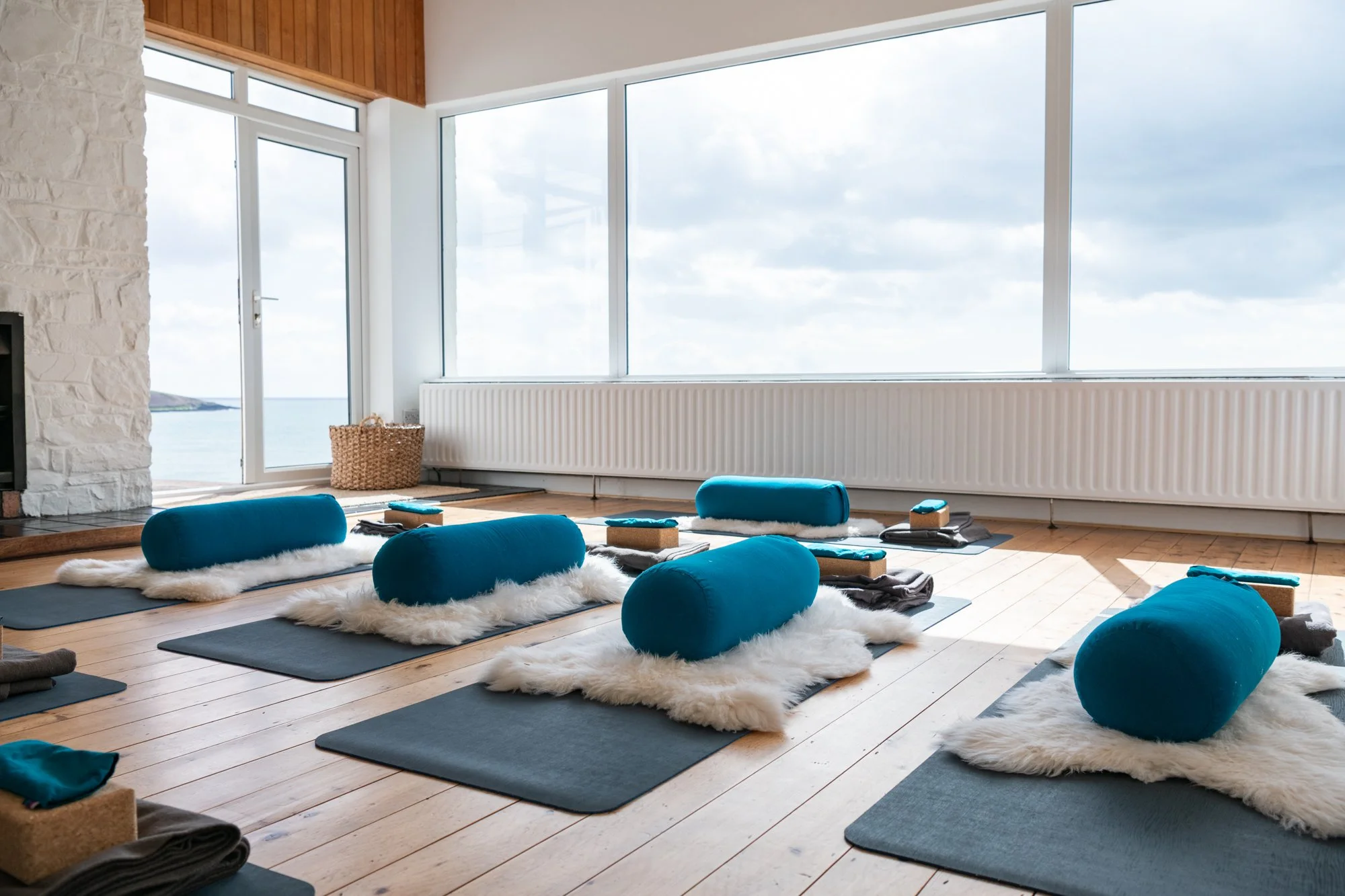 Yoga studio with large windows overlooking the ocean, featuring mats, turquoise bolsters, and fluffy white blankets on wooden floors.