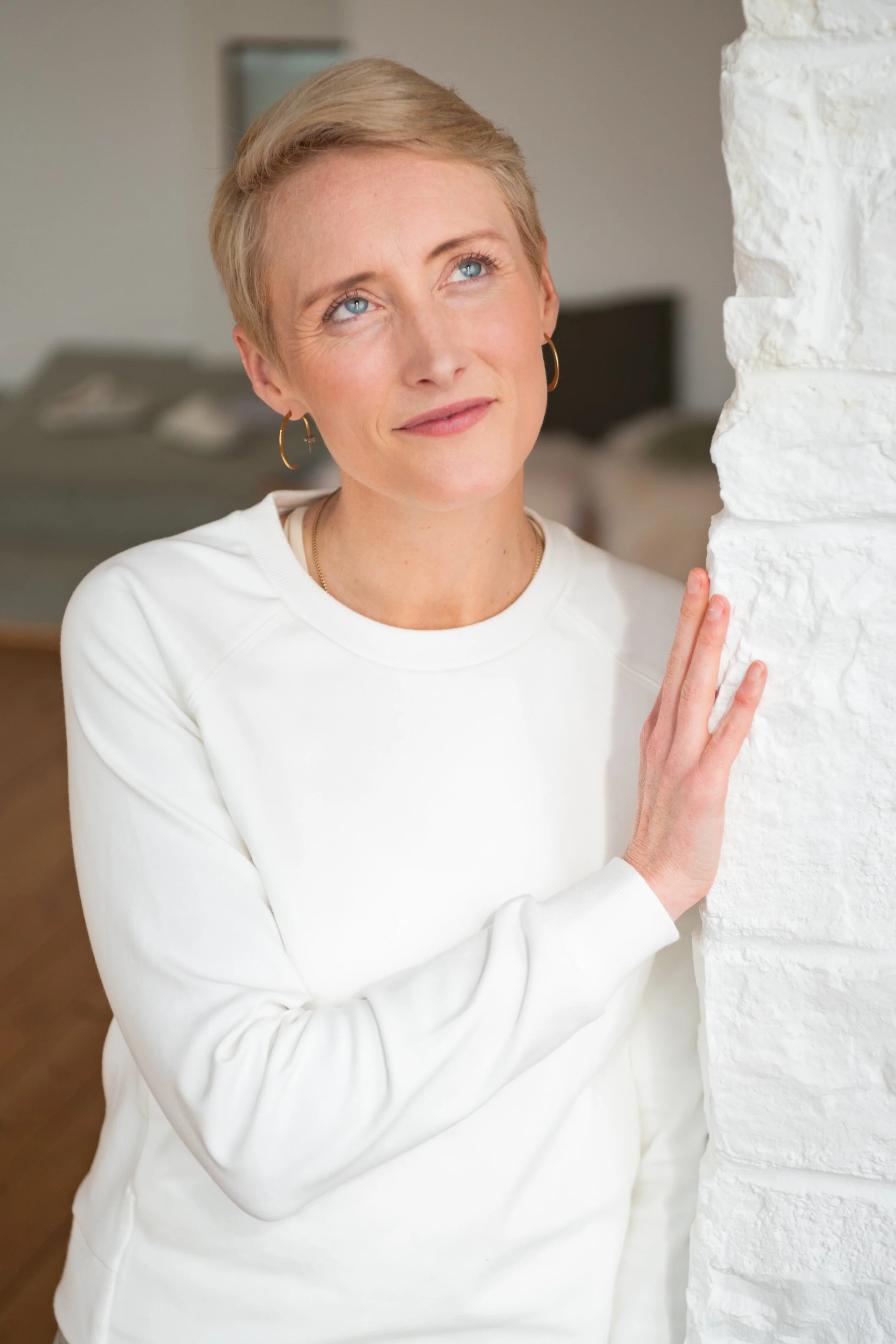 Person with short blonde hair wearing a white sweatshirt, looking thoughtful while leaning against a white brick wall.