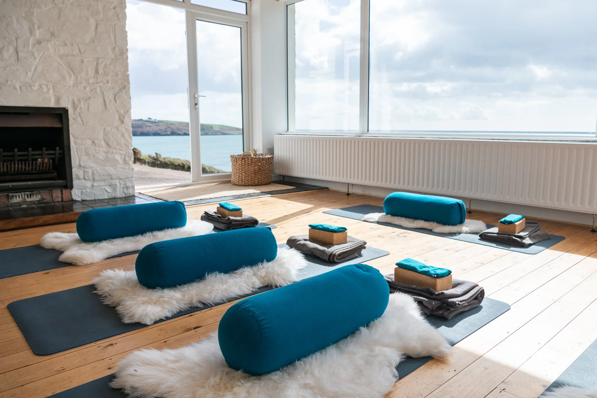 Yoga studio with wooden floor, blue bolsters on fluffy mats, and large windows offering a sea view.