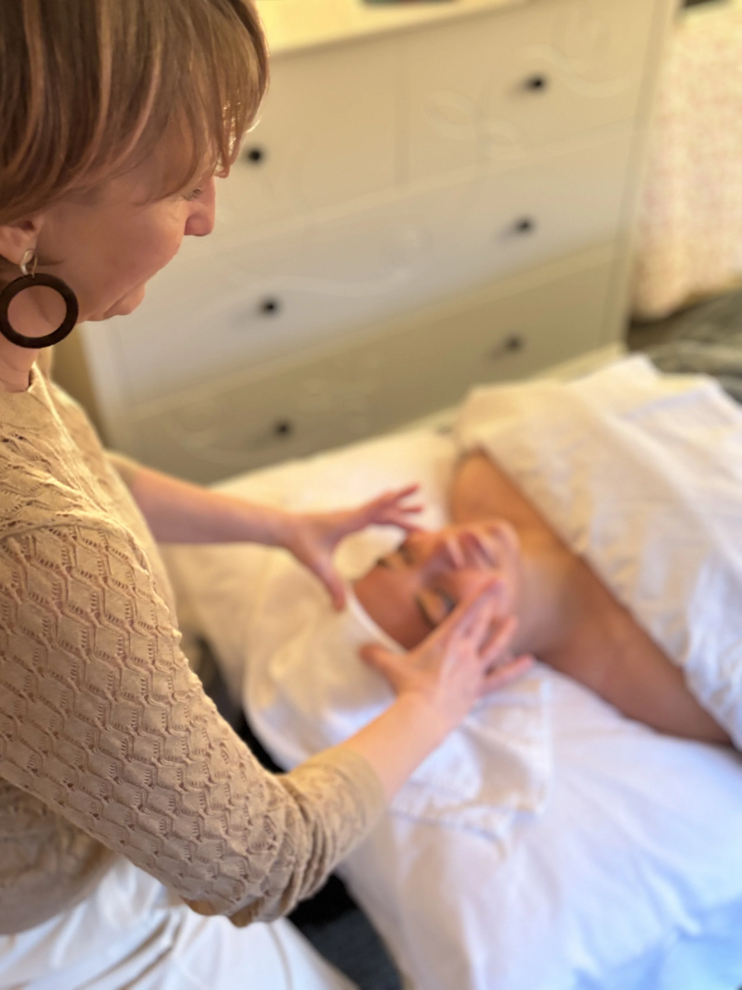 Lucie of Laurea Studio Victoria providing facial reflexology to a person lying on a bed, with the woman's hands on the person's thighs, in a softly lit room.