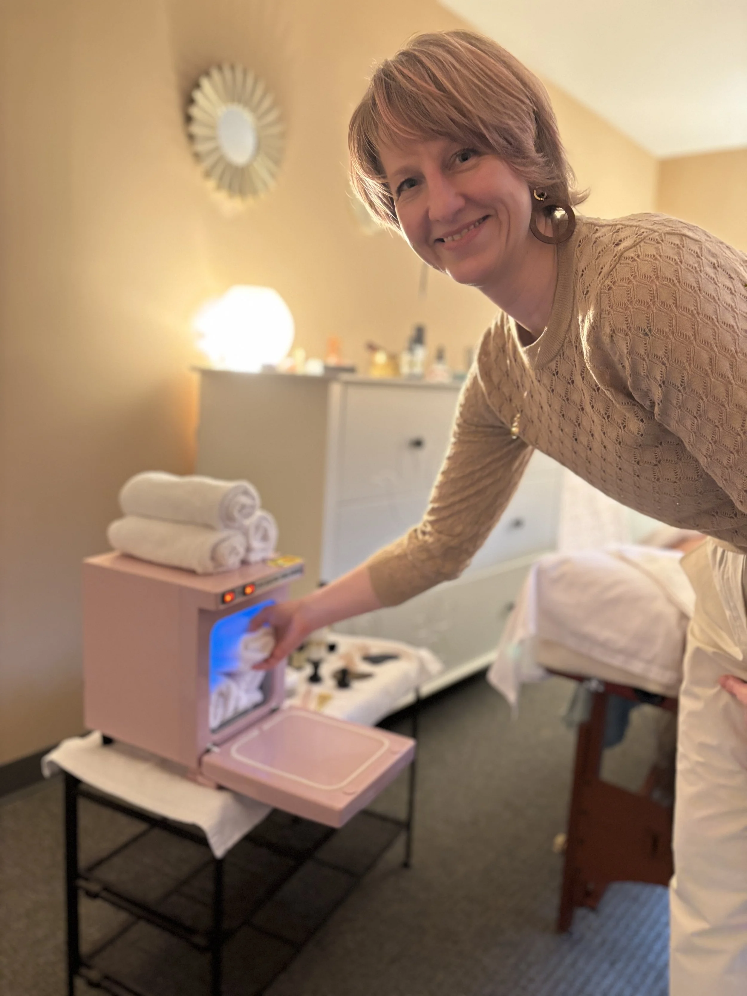 Lucie of Laurea Studi in a beige sweater is placing a white towel on a pink towel warmer in a room with beige walls, illuminated by warm lighting. There are neatly rolled towels on top of the towel warmer and a dresser with bottles in the background.