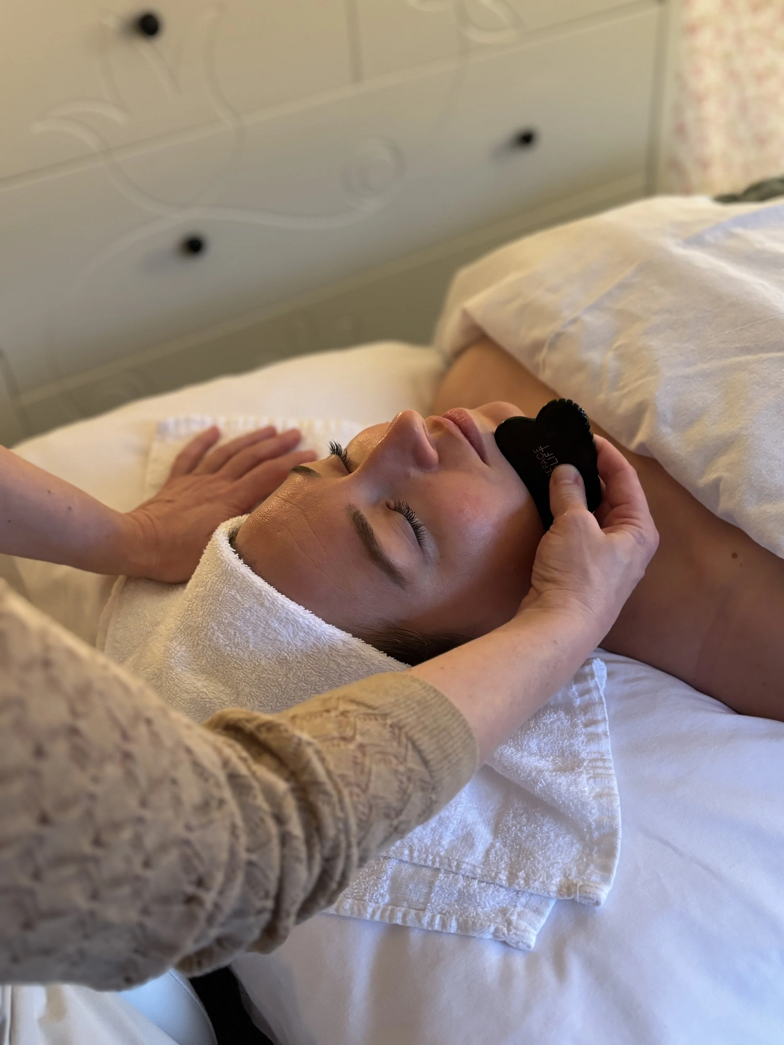 A woman lying on a bed with her eyes closed, receiving a facial reflexology from Lucie at Laurea Studio Victoria. The woman has a towel wrapped around her head and is resting on a white towel.