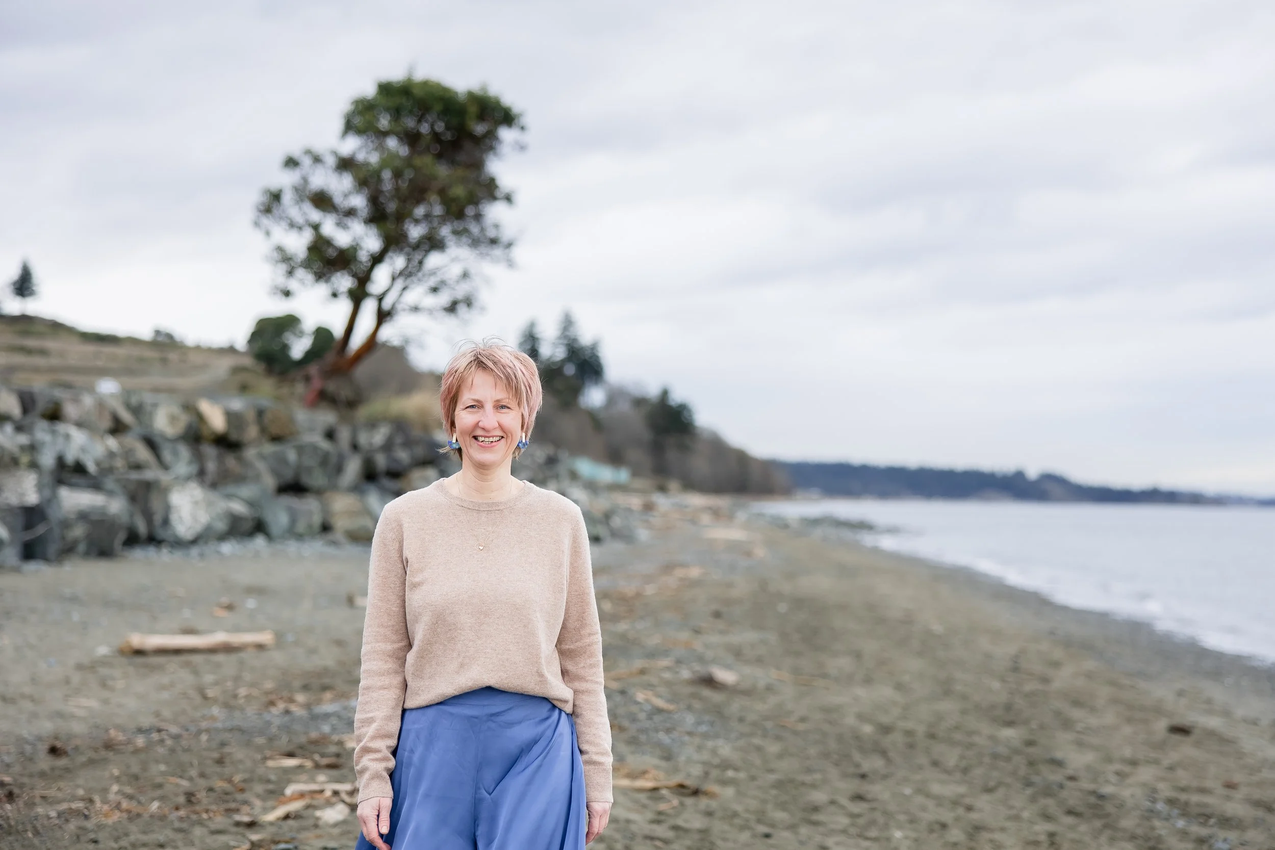 Lucie, founder of Laurea Studio, standing on a rocky beach near the water, with trees and cloudy sky in the background.