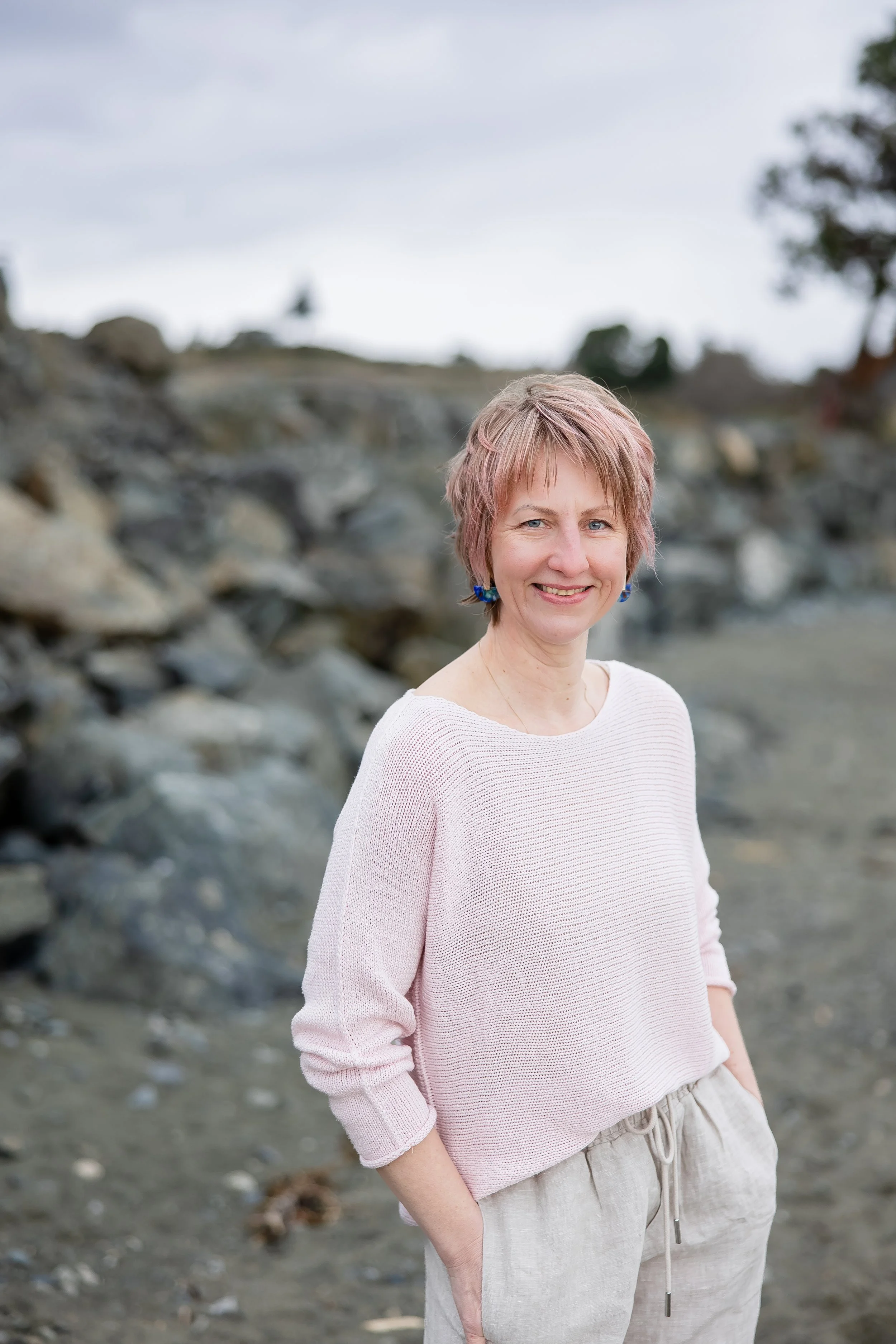 Lucie, facial reflexologist at Laurea Studio,  smiling outdoors on a rocky beach with a cloudy sky and trees in the background.