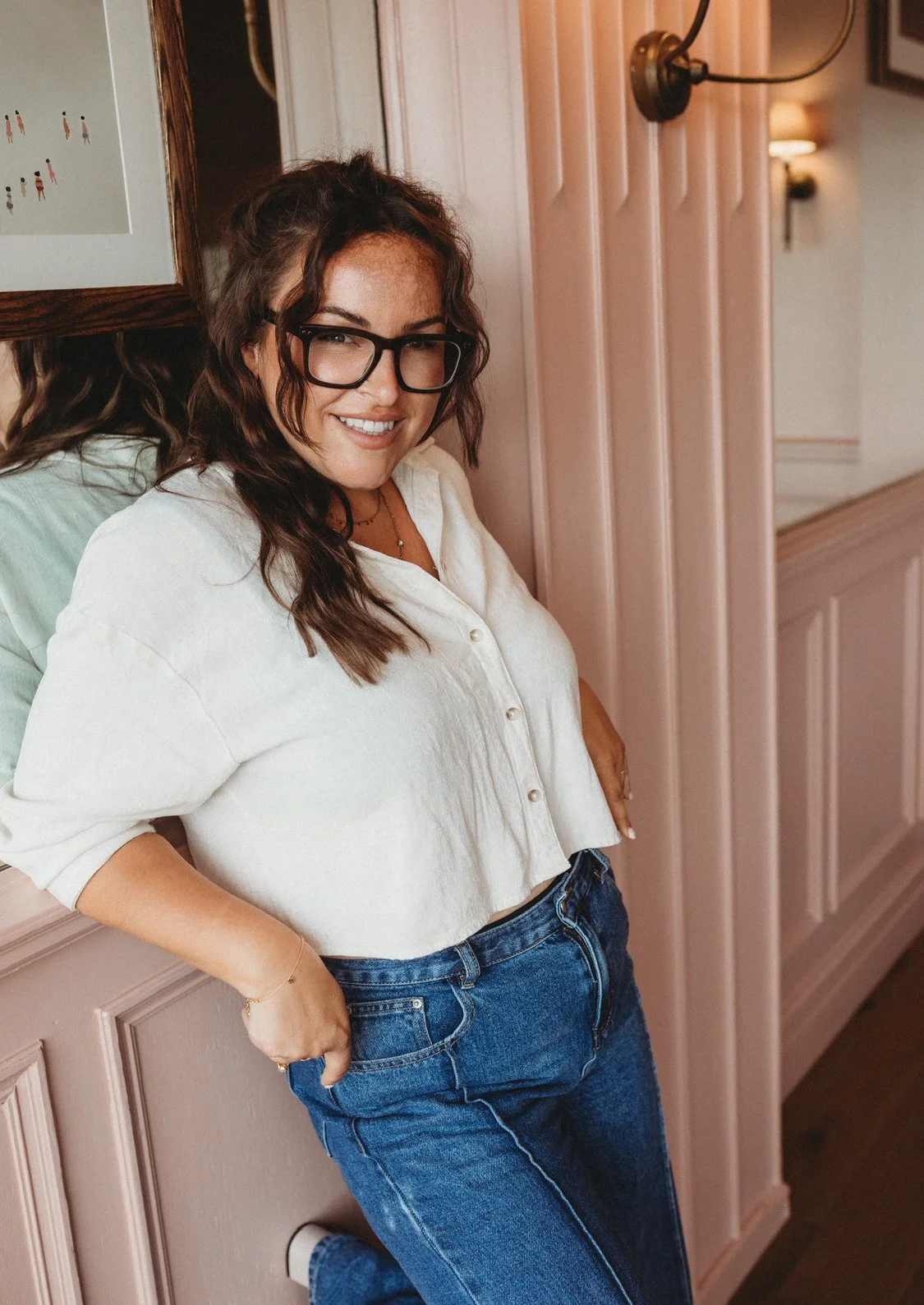 A woman with dark wavy hair, glasses, and a white shirt, smiling and leaning against a pink wall with his hand in her pocket.
