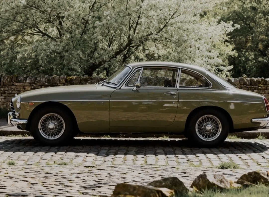 A vintage olive-green coupe car parked on a cobblestone street with a stone wall and trees in the background.