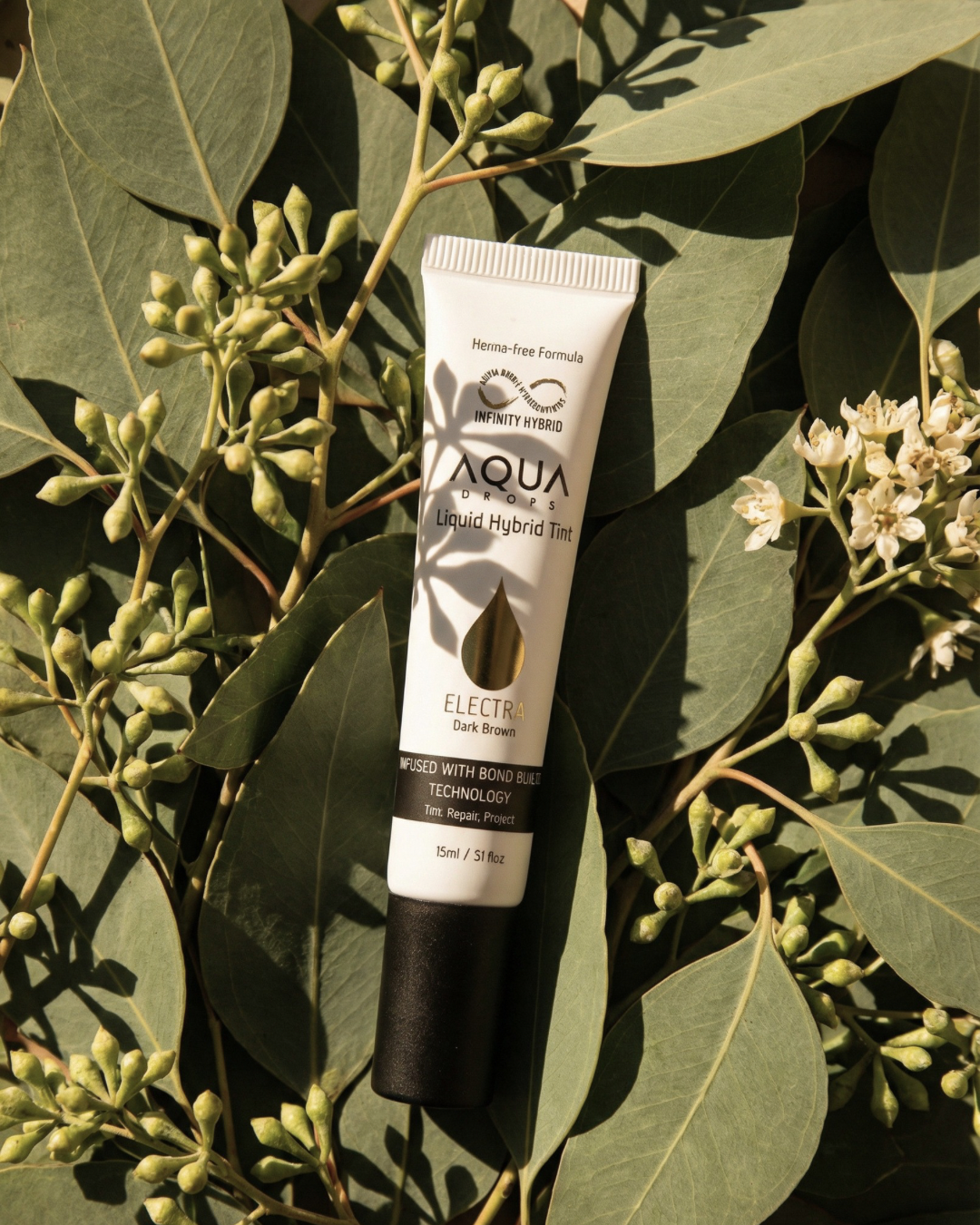 A white tube of Aqua Drops Liquid Hybrid Tint, Electra Dark Brown, placed on green eucalyptus leaves and flower buds.