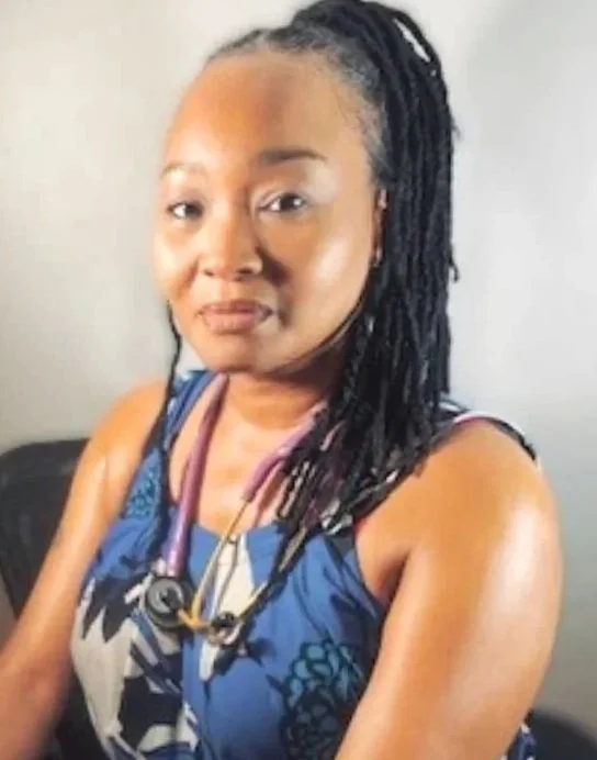 A woman with braided hair wearing a blue floral top and a stethoscope around her neck, sitting indoors.