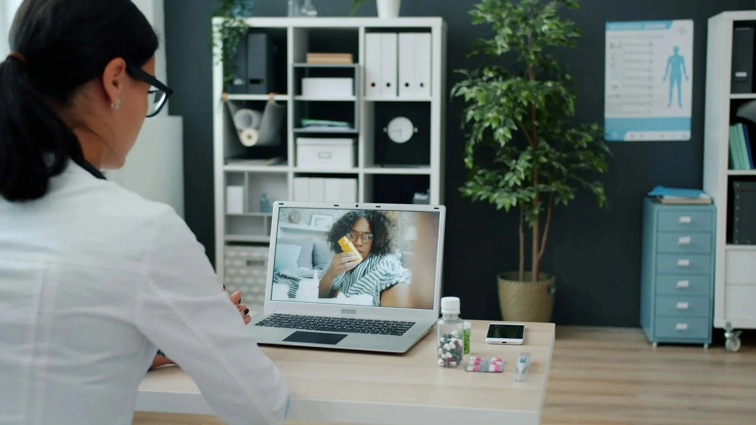 A woman is looking at a woman on a video call on her laptop, with medication and a phone on the desk in front of her. The room has shelving with office supplies, a potted plant, and a medical poster on the wall.