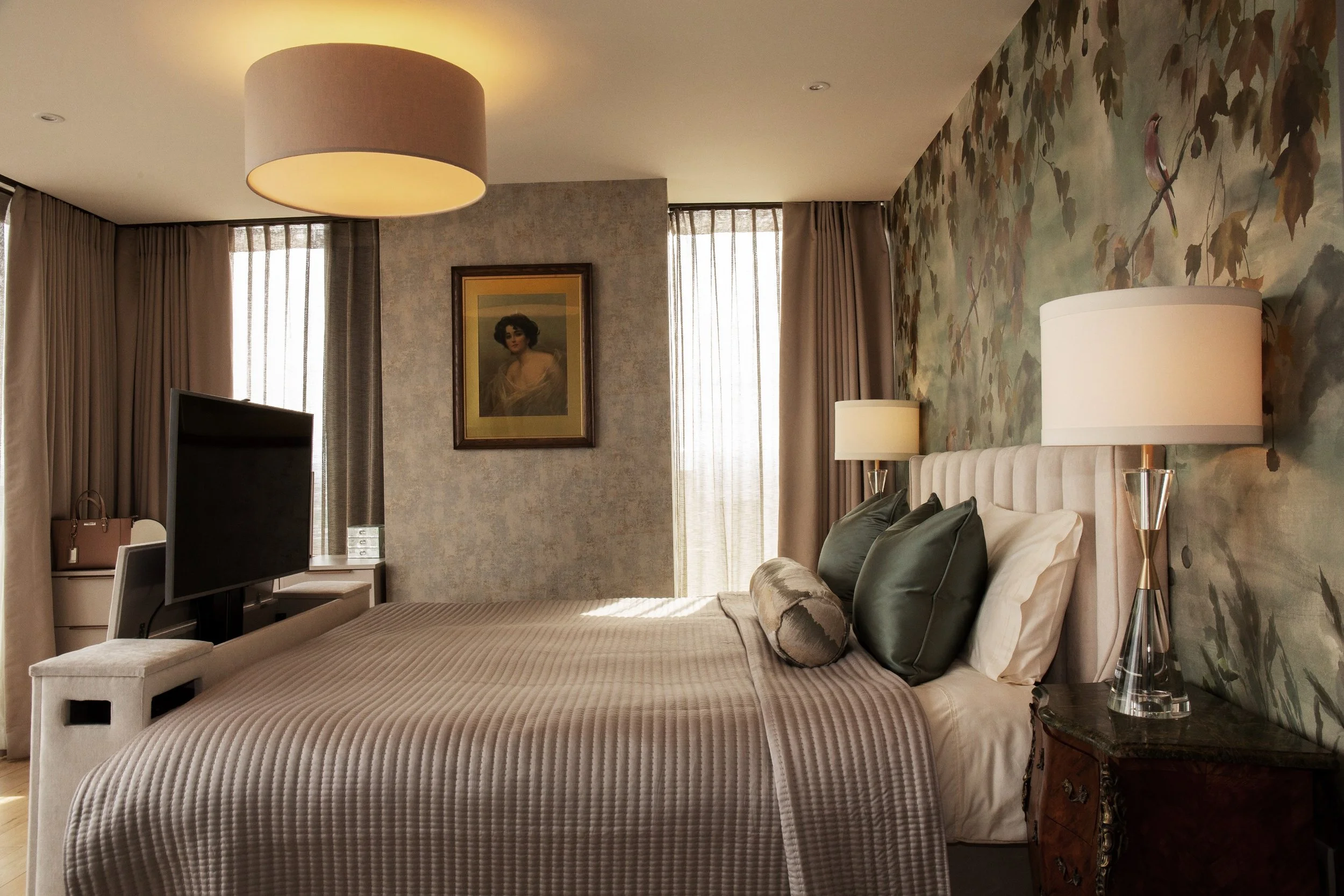 A neatly made bed with green pillows and a textured beige bedspread in a hotel room, with a floral accent wall behind the headboard, flanked by two lamps, and a flat-screen TV on a stand at the foot of the bed. A framed portrait hangs on the wall, with beige curtains on windows allowing natural light.