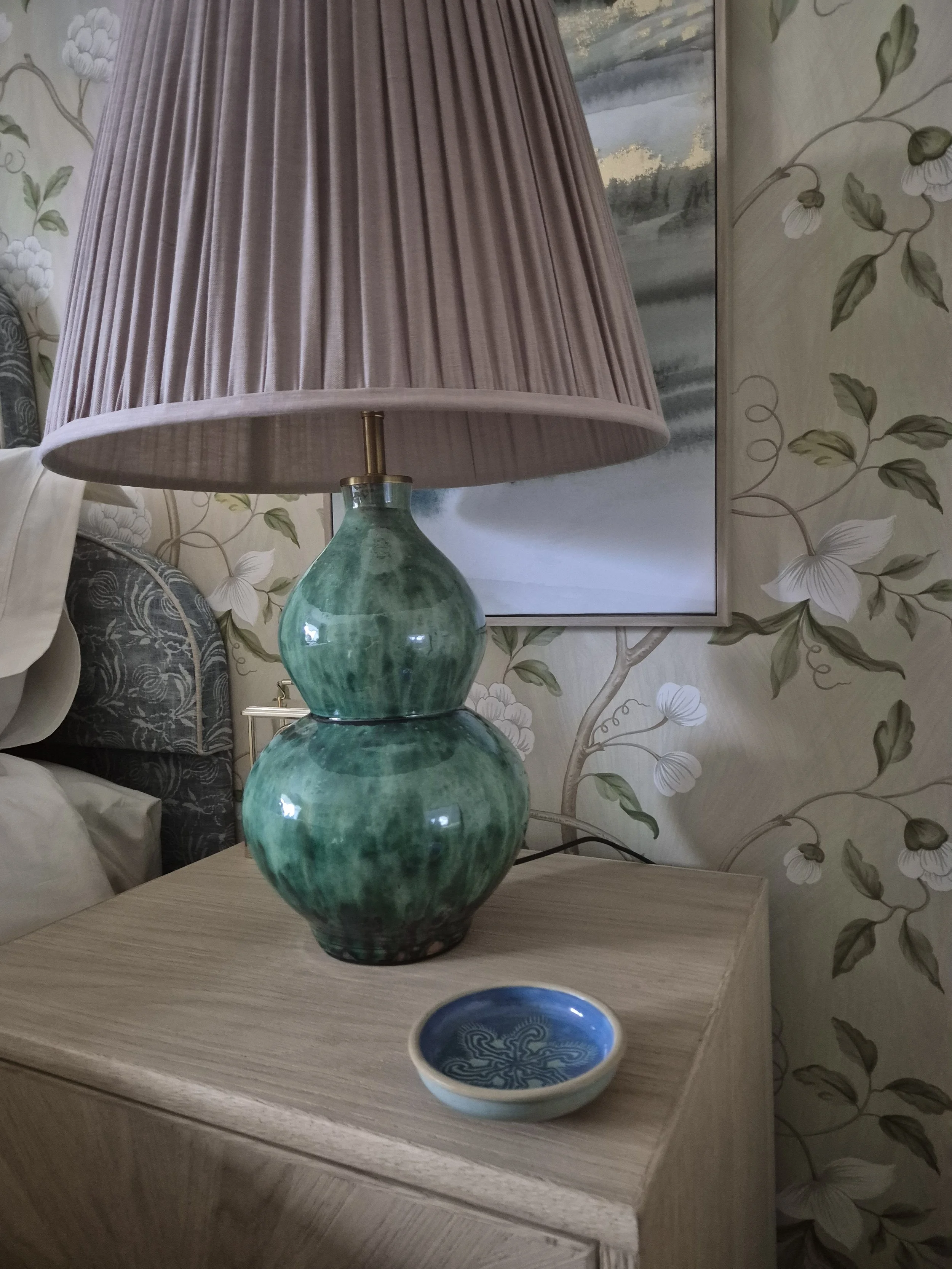 A green double-gourd shaped table lamp with a pink pleated lampshade on a light wood nightstand, with floral wallpaper and a framed painting in the background.