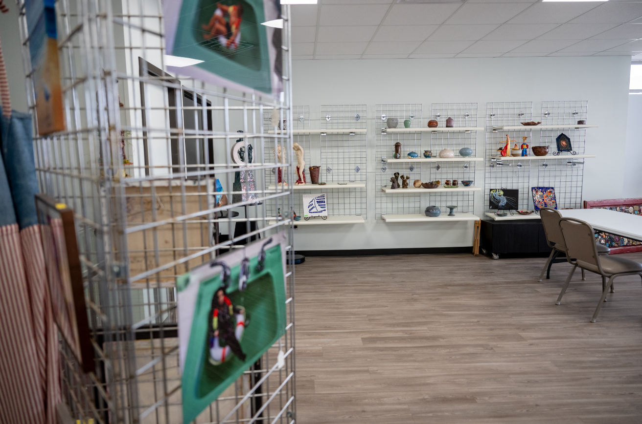 Art gallery interior with wire mesh display, artwork on shelves, and photos on metal grids. Wooden floor, chairs, and table visible.