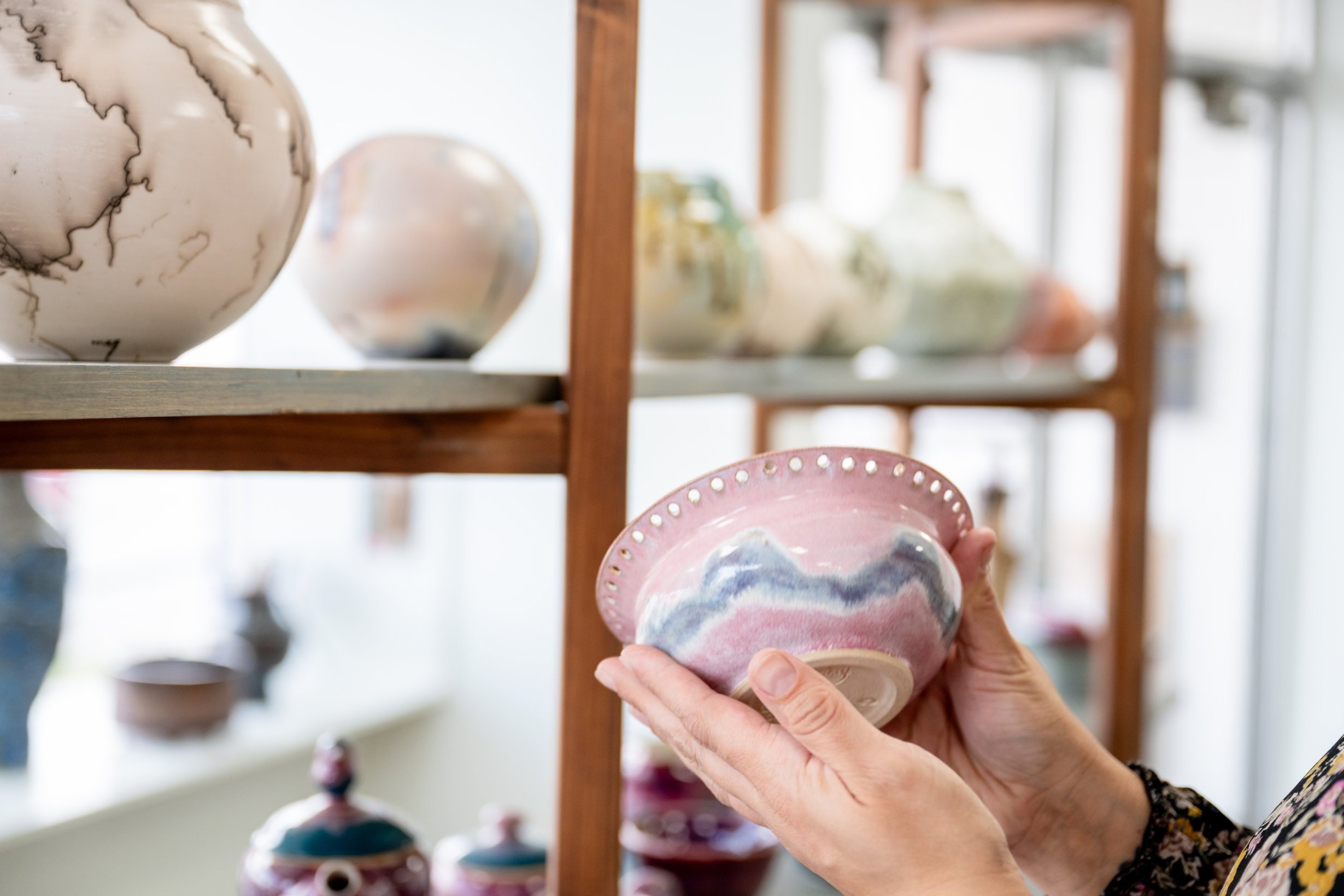 A person holding a handcrafted ceramic bowl with pink and blue glaze, surrounded by various pottery items on wooden shelves.