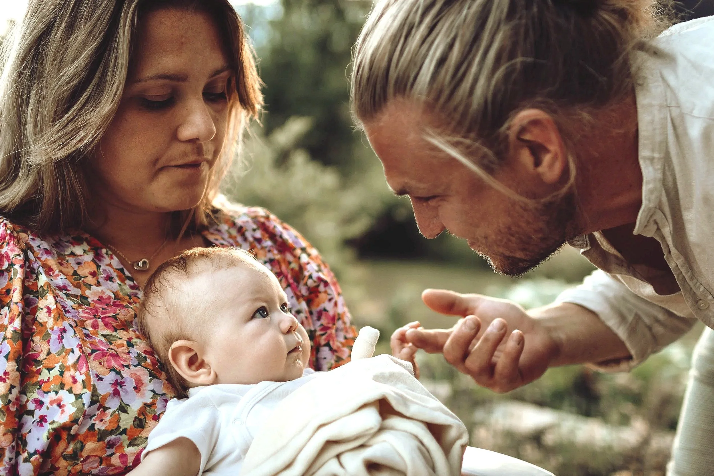 Ett barn som ligger i en vuxen kvinnas knä, medan en man lutar sig nära och tittar på barnet, i ett naturskönt utomhusmiljö.