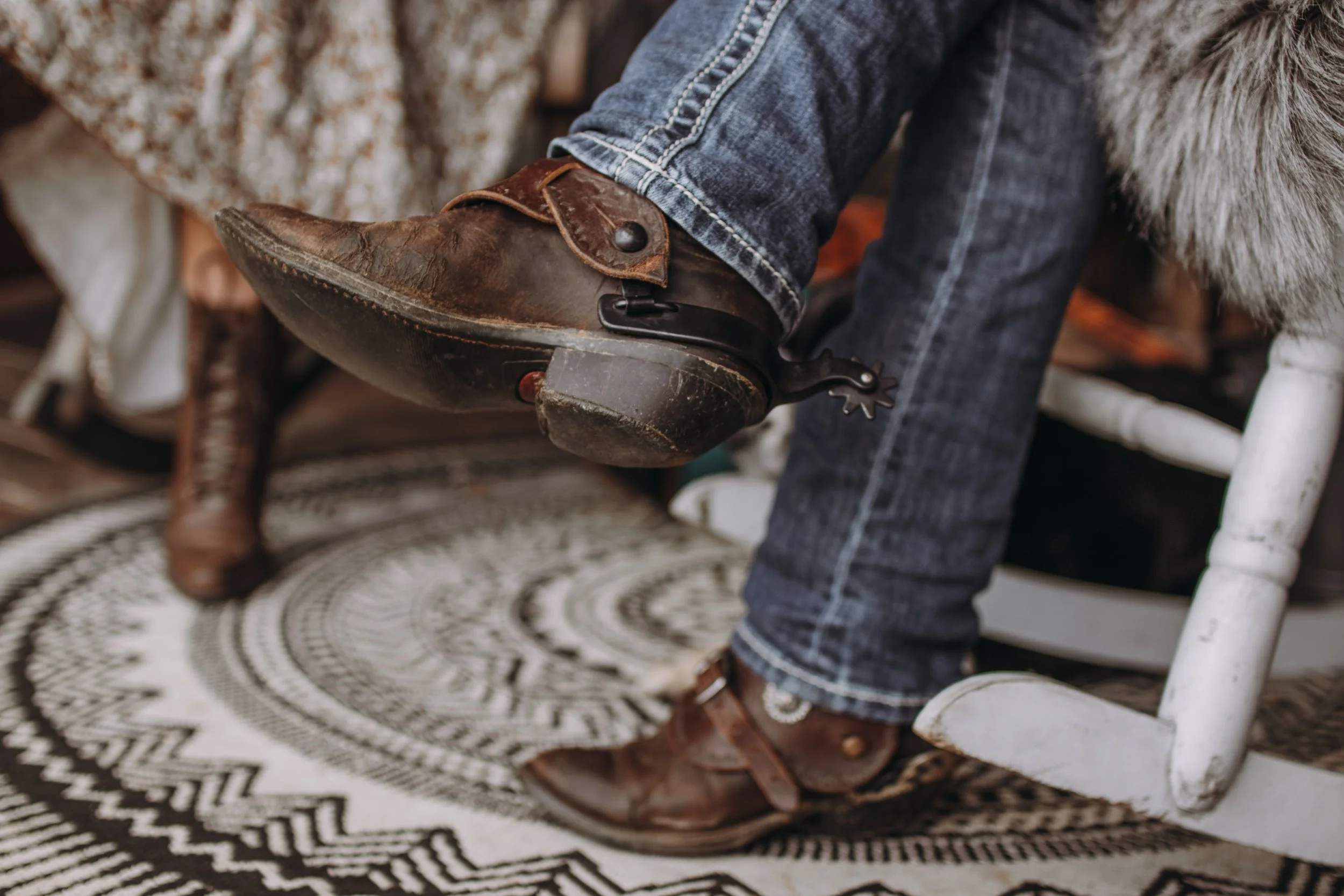 En person som sitter på en vit trästol, klädd i jeans och skor, som kopplar en vintage stövel som är lagad med en protes.