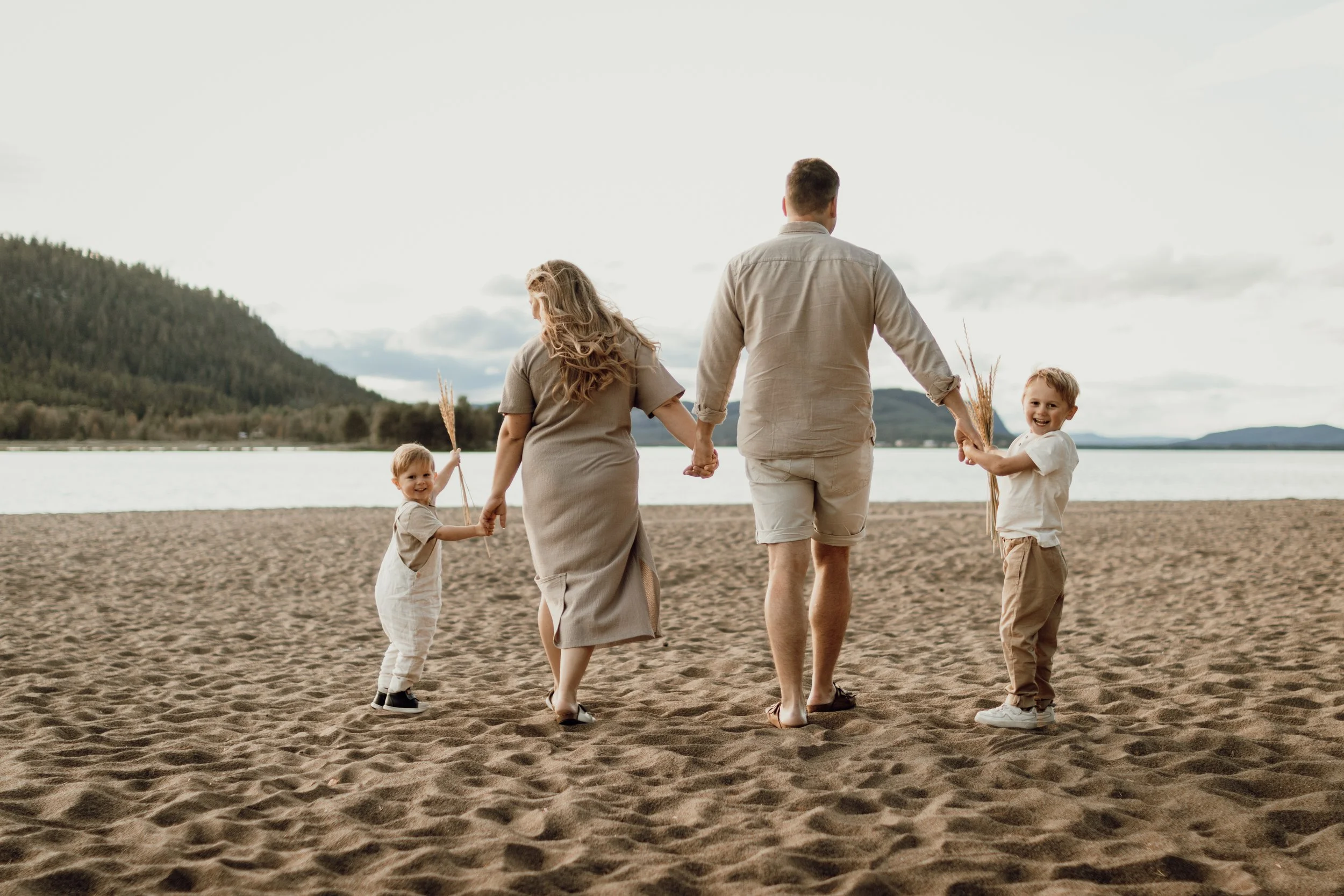 En familj av fyra som går på en sandstrand vid en sjö eller hav, håller handen och bär gräspinnar, med ett landskap av kullar och vatten i bakgrunden, under en molnig himmel.