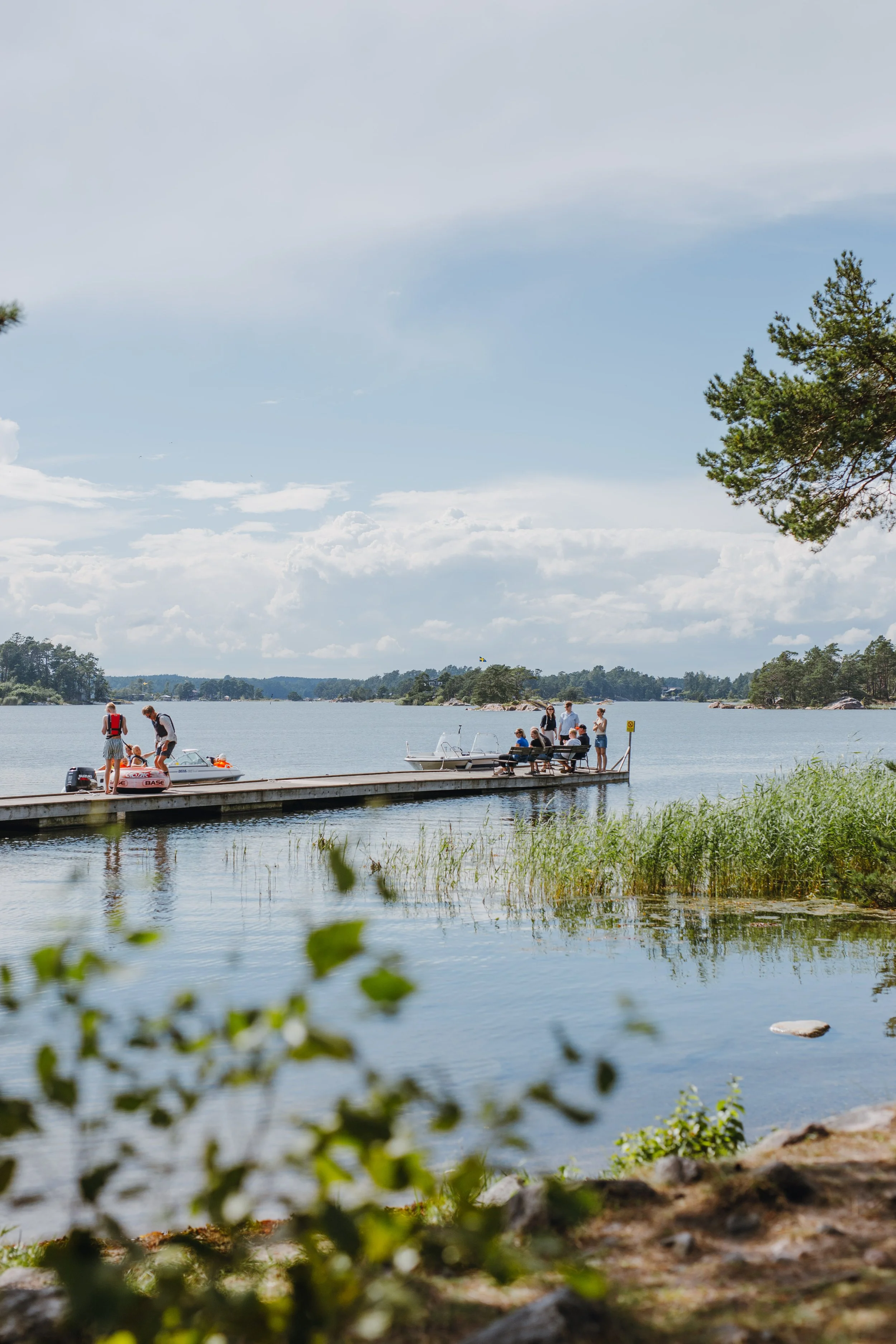 Personer på en brygga vid en sjö, några sitter och andra står, med båtar i närheten och grönska runt omkring.