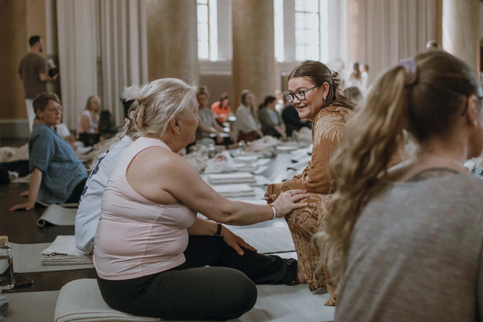 Två kvinnor pratar och ler i ett rum med många andra människor som sitter vid ett långbord, vilket tyder på en social eller formell sammankomst.