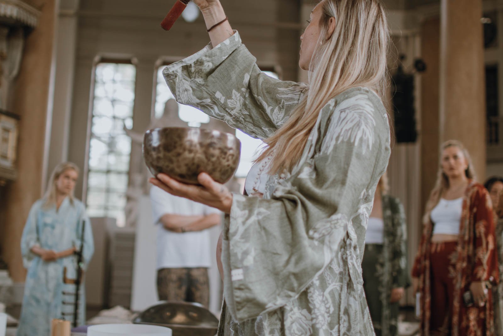 En kvinna som håller en rökelsekopp i ena handen och en sked i den andra, omgiven av flera andra kvinnor i ett rum med höga fönster.