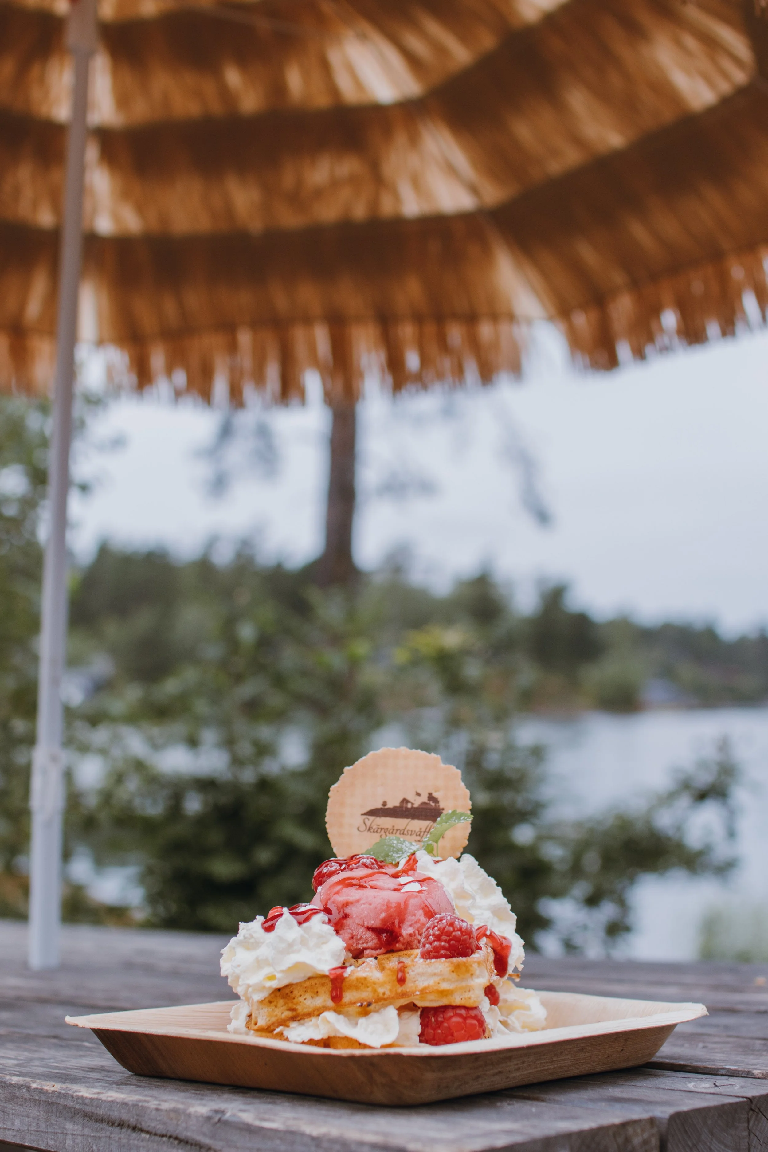 En dessert med våffla, grädde, hallon och jordgubbar på en träbricka utomhus under ett stråtak, med vatten och träd i bakgrunden.