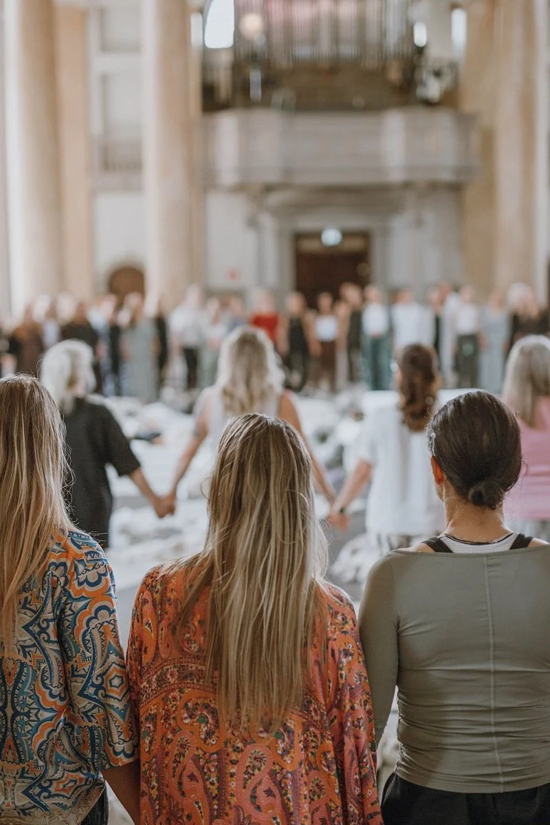 Kvinnor i St Ericsson Hallen som står i två större ringar och håller händerna på varandra, närbild på tre av dem med ryggen mot kameran