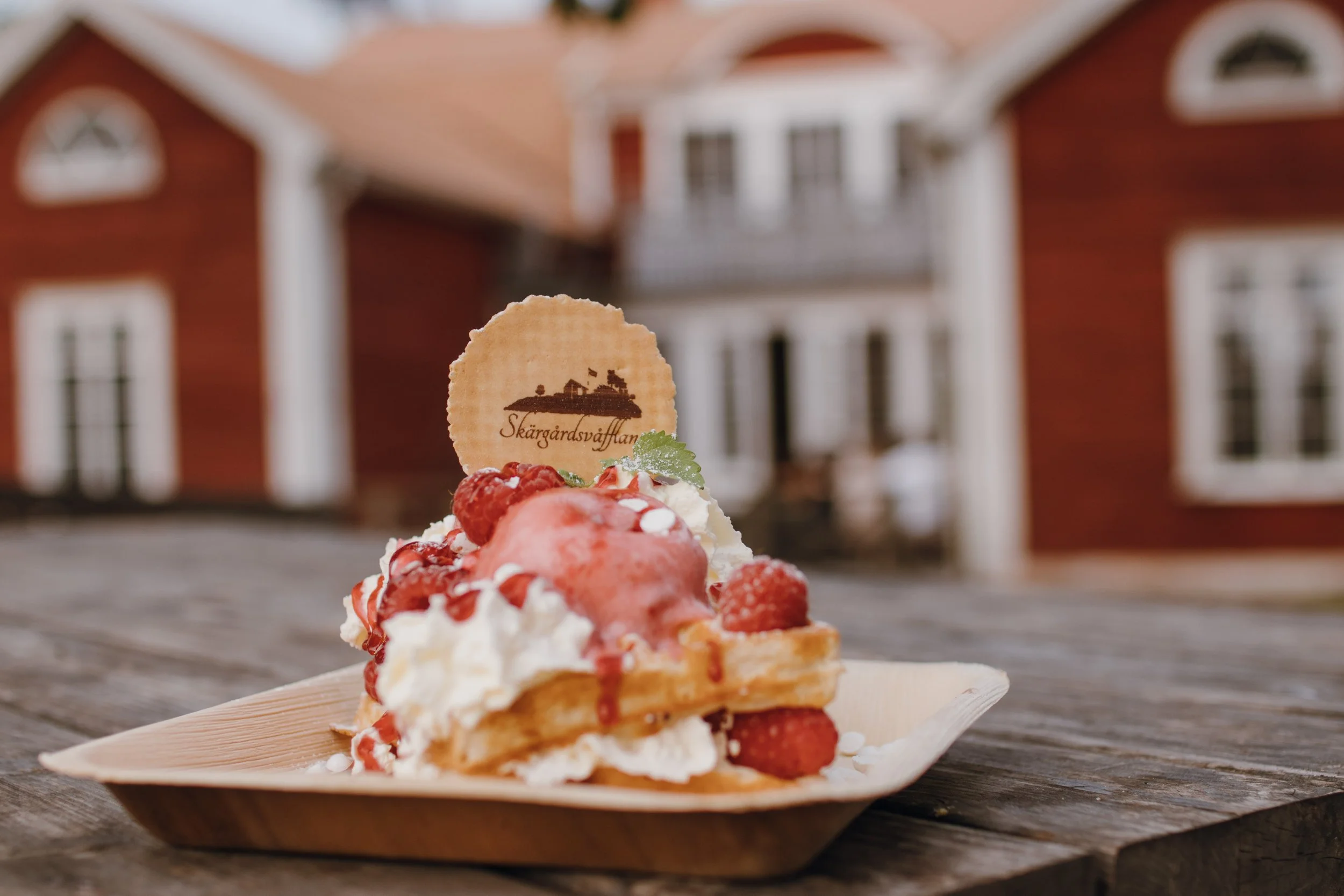 En bit av en jordgubbstårta med vispgrädde, jordgubbar och ett rundkaka med texten 'Skärgårdsvåfflan'.