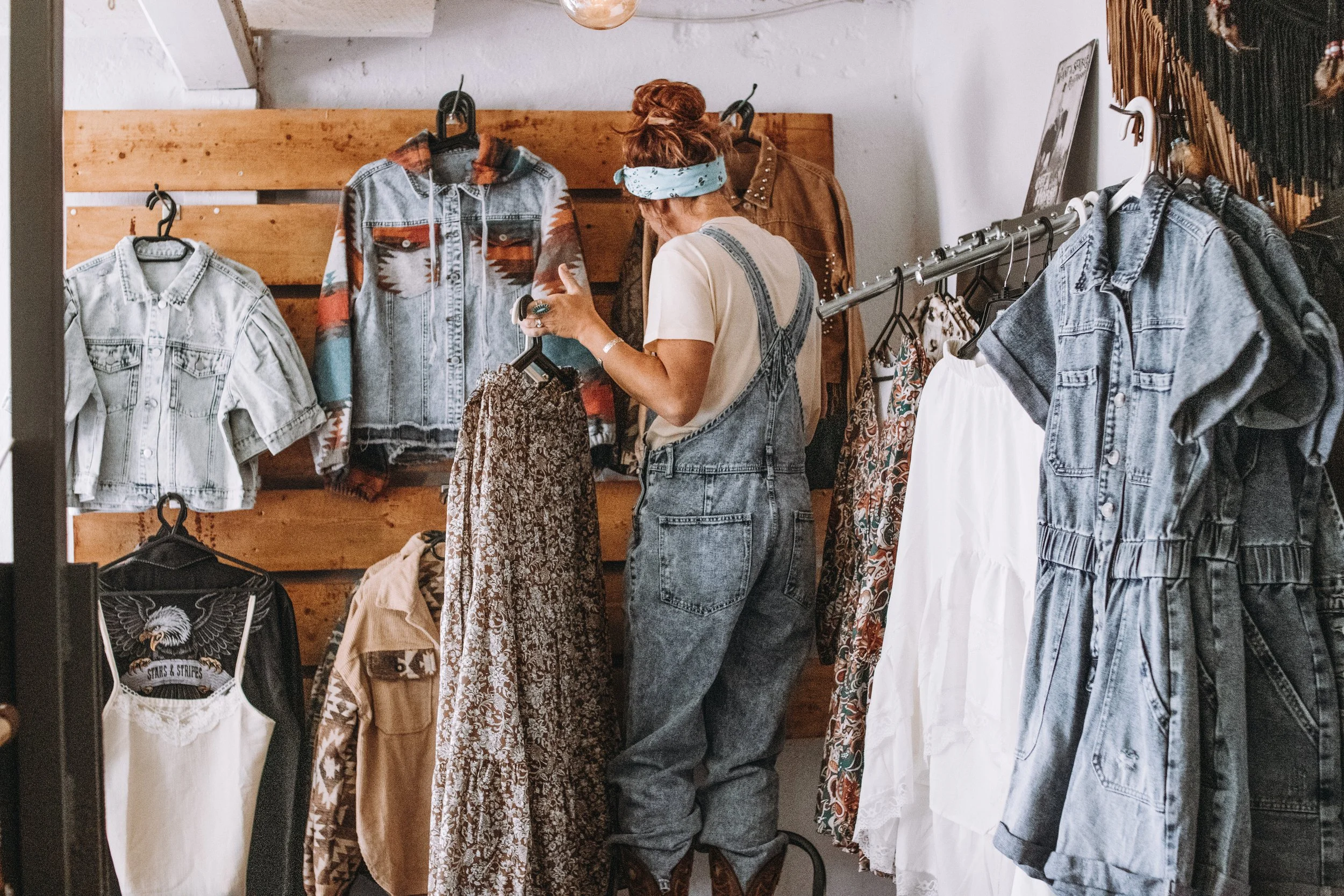 Kvinna som provar kläder i en secondhandbutik, omgiven av olika plagg av denim, blommiga och vita tyger.