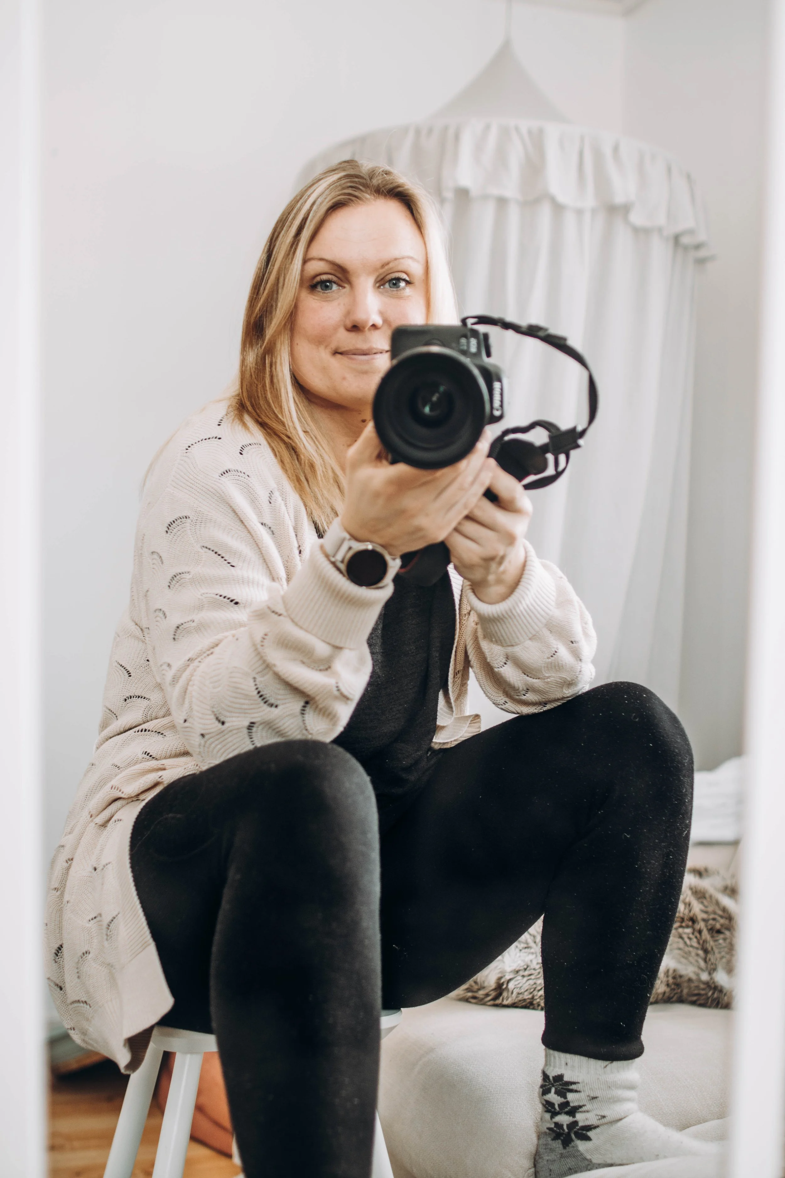 Kvinna sitter på en pall och har fötterna på ett fotstöd med kameran synlig i handen riktad mot åskådaren