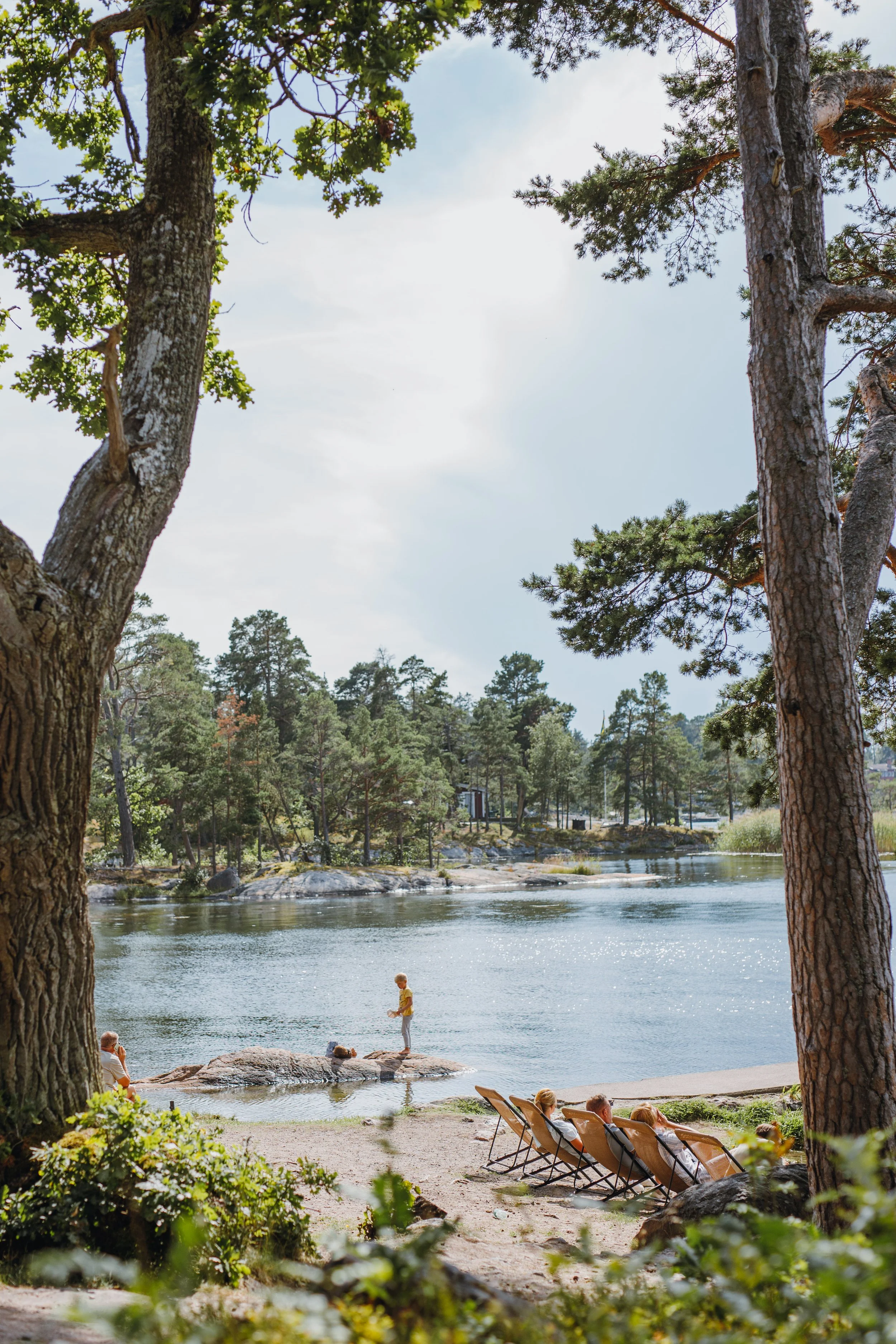 Barn som leker på en klippa vid en sjö, medan vuxna ligger och solar på stranden, omgiven av träd.