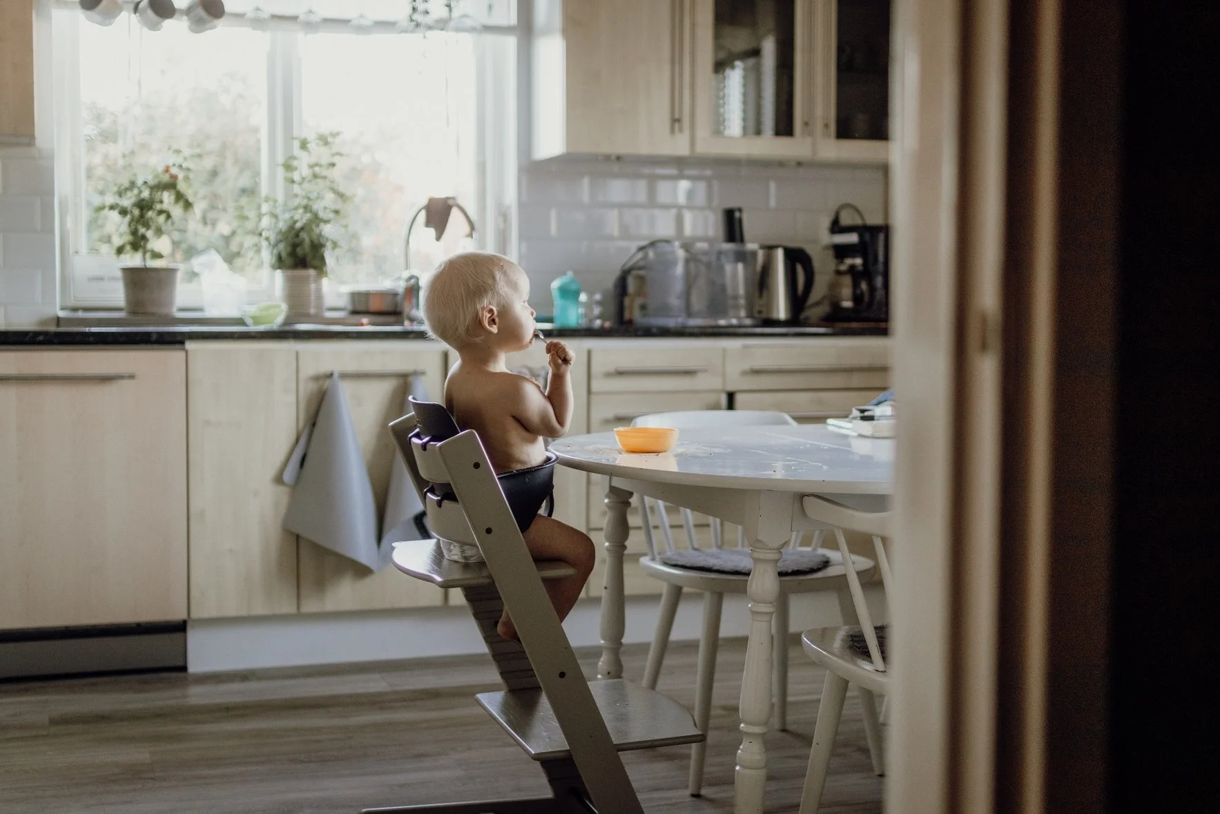 2 åring sitter ensam vid köksbordet och äter, bilden är tagen snett bakifrån en bit ifrån