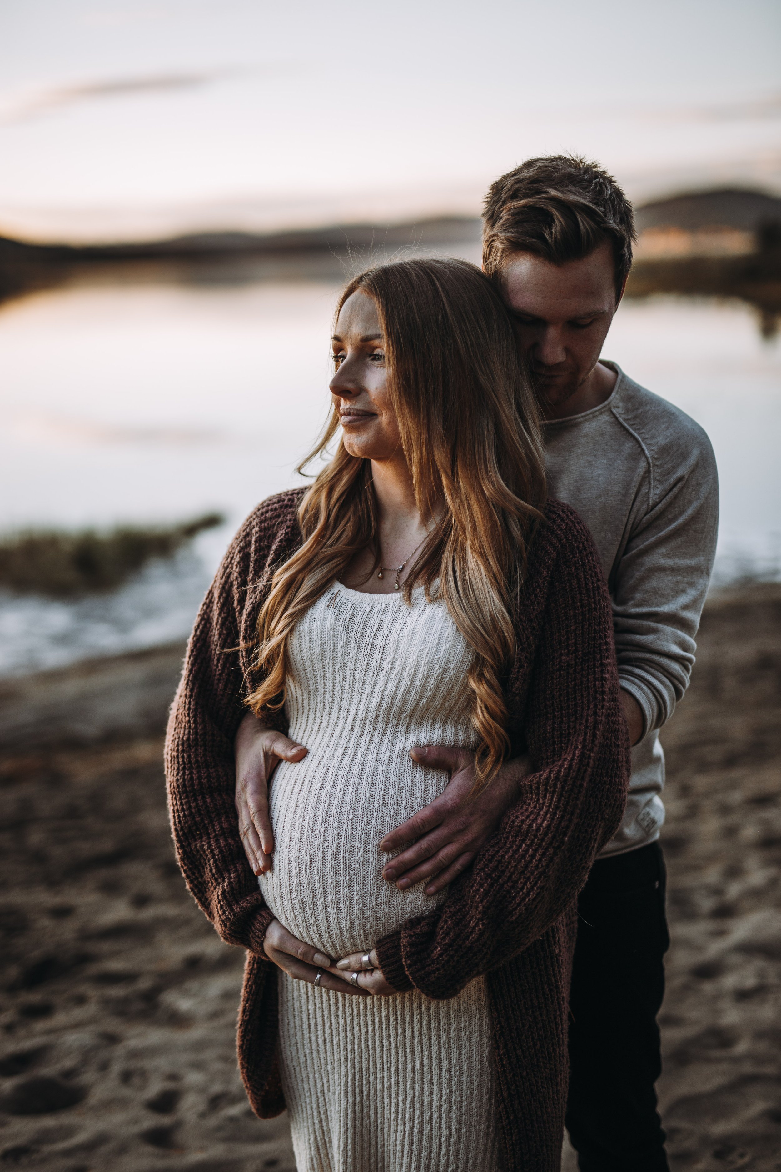 En gravid kvinna med långt, lockigt hår står på stranden, omgiven av en man som håller runt hennes mage och omsluter henne med händerna. Solnedgången skiner i bakgrunden.