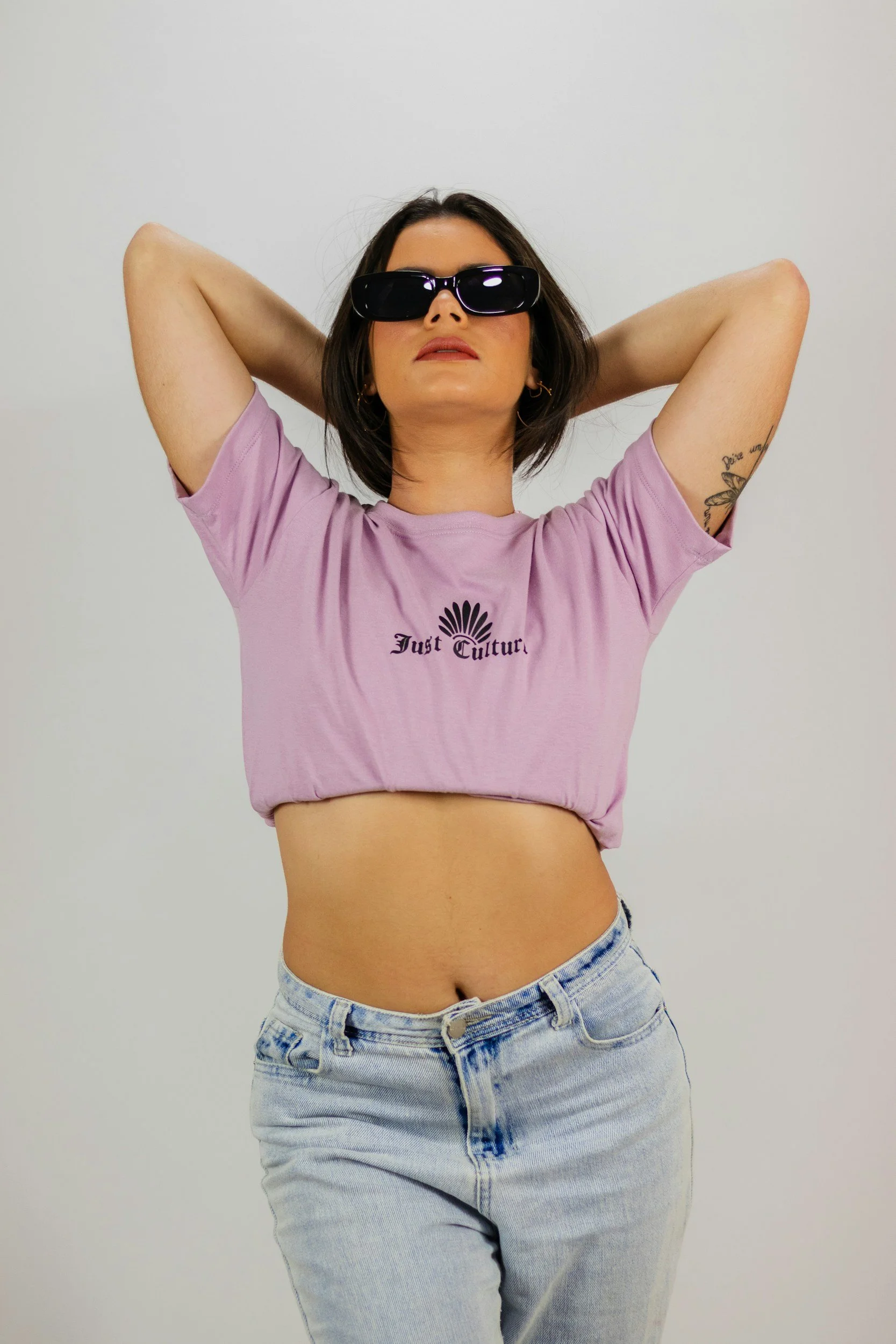Femme portant un t-shirt violet et des lunettes de soleil, bras levés derrière la tête.
