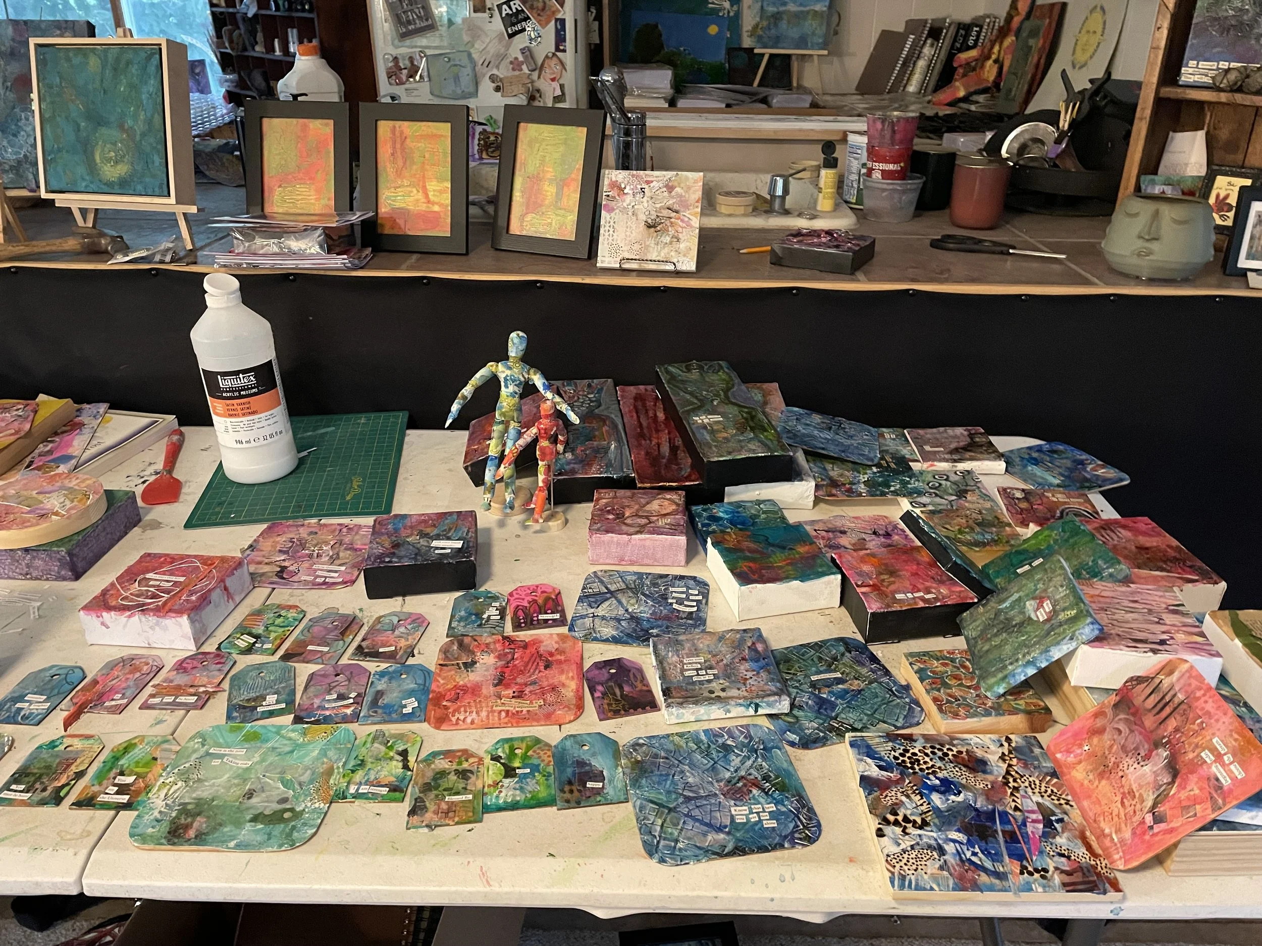 A cluttered art studio workspace with colorful abstract paintings, small canvases, painted figurines, art supplies, and framed artwork on the table and shelves.