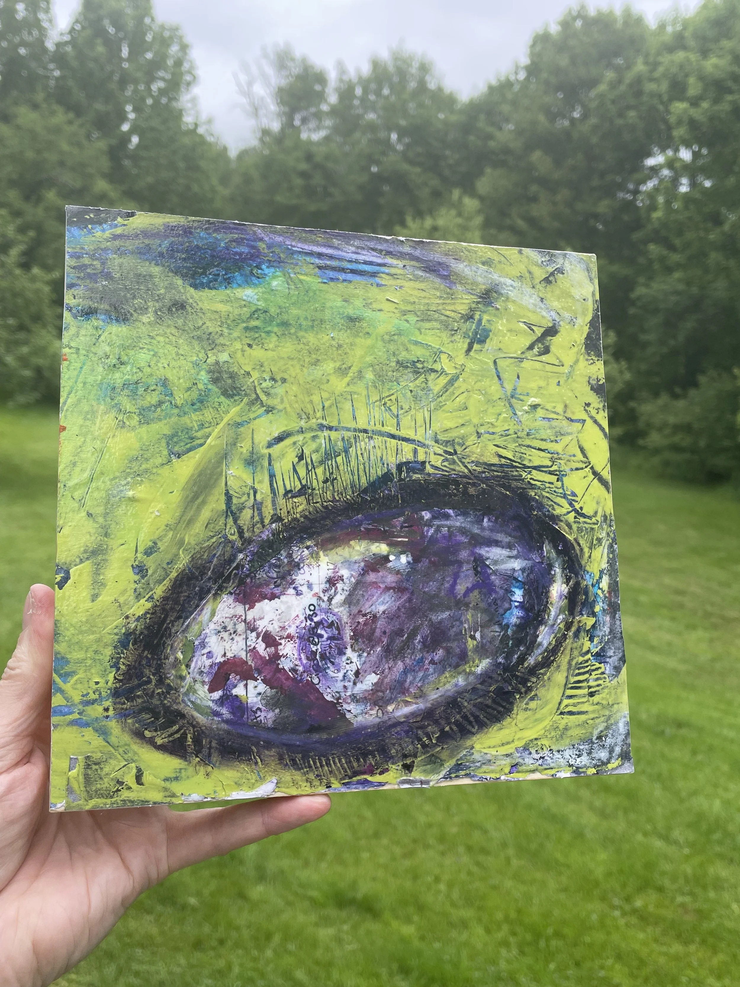 Hand holding a colorful abstract painting with a large eye shape, set outdoors with green grass and trees in the background.