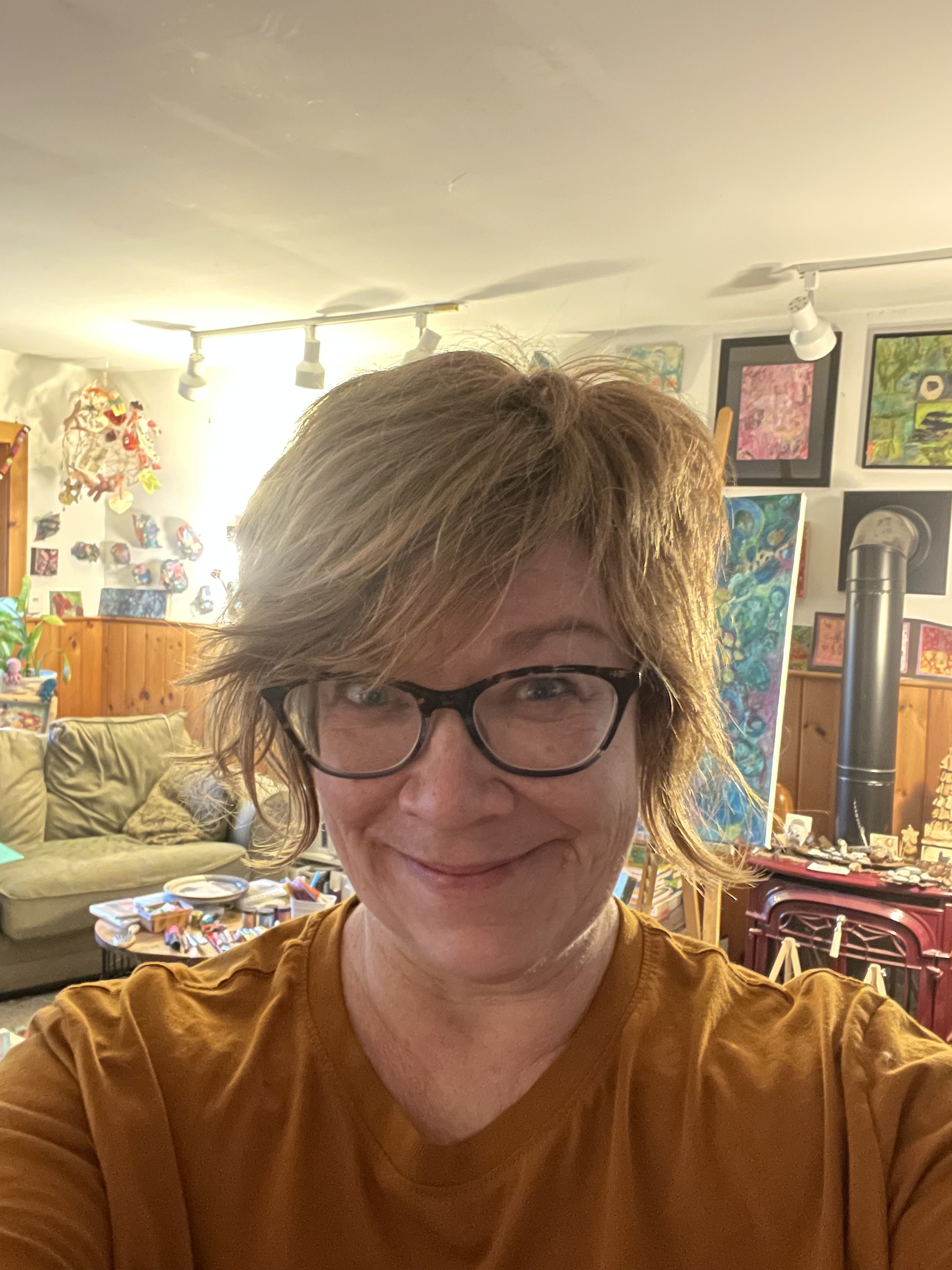 A woman with short, tousled hair and glasses smiling at the camera inside a cozy, art-filled room with paintings on the walls, a wood stove, and various items on tables.