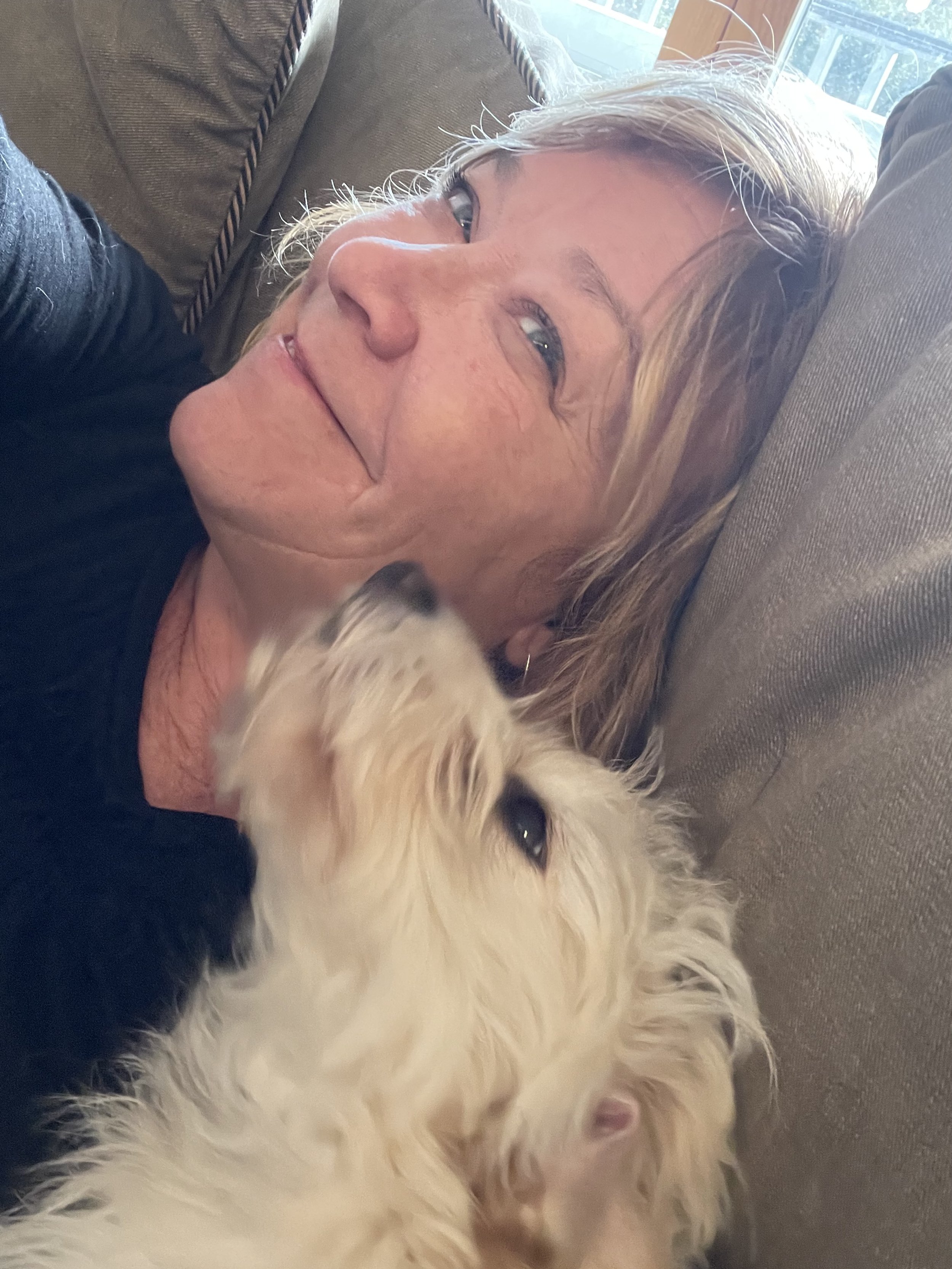 An older woman with light hair lying on a couch with a black and white dog resting its head on her shoulder.