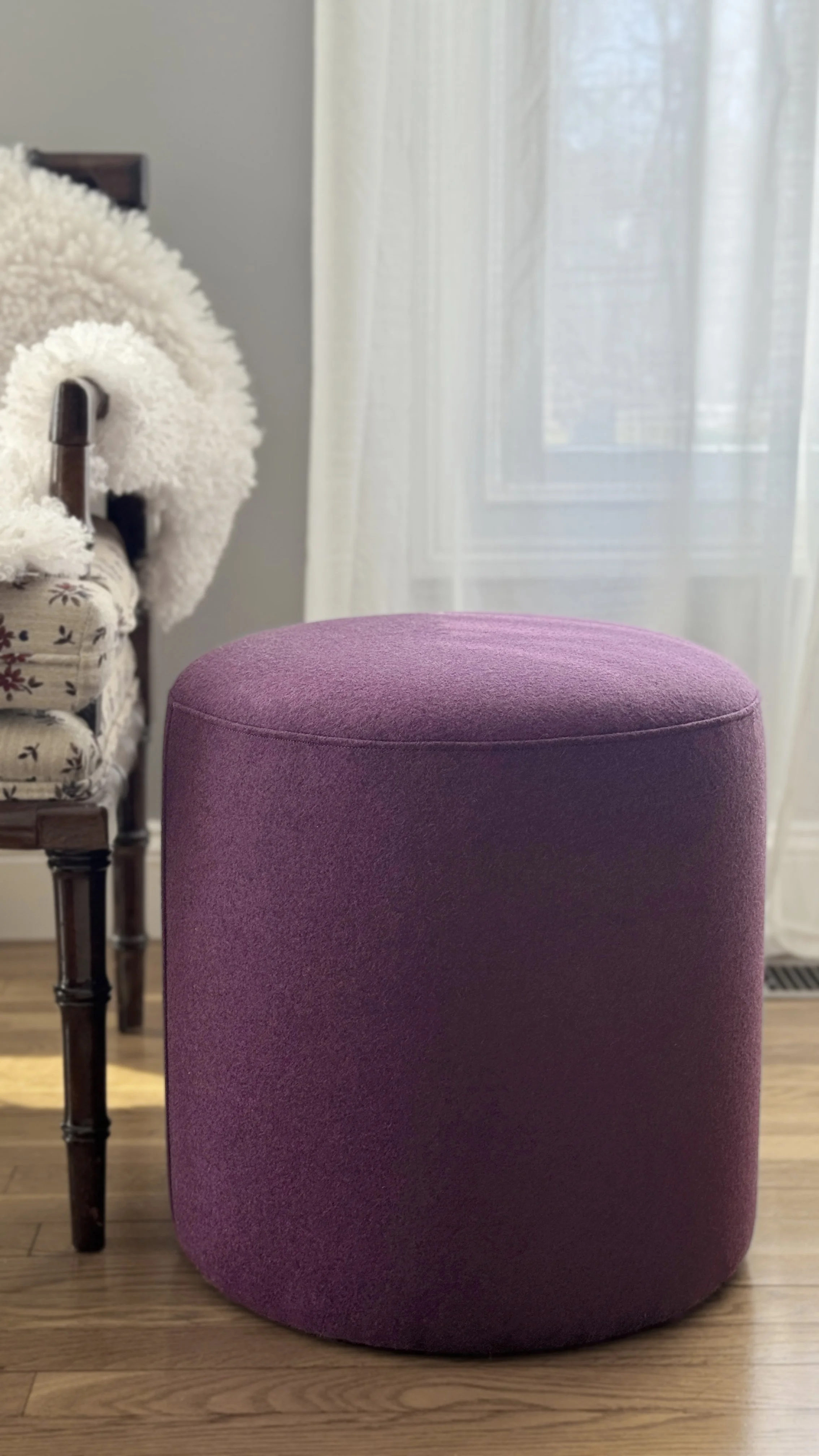 A round, purple upholstered ottoman in front of a wooden floor, with a white curtain and a window in the background. To the left, there is a wooden chair with a floral cushion and a white fluffy throw draped over it.