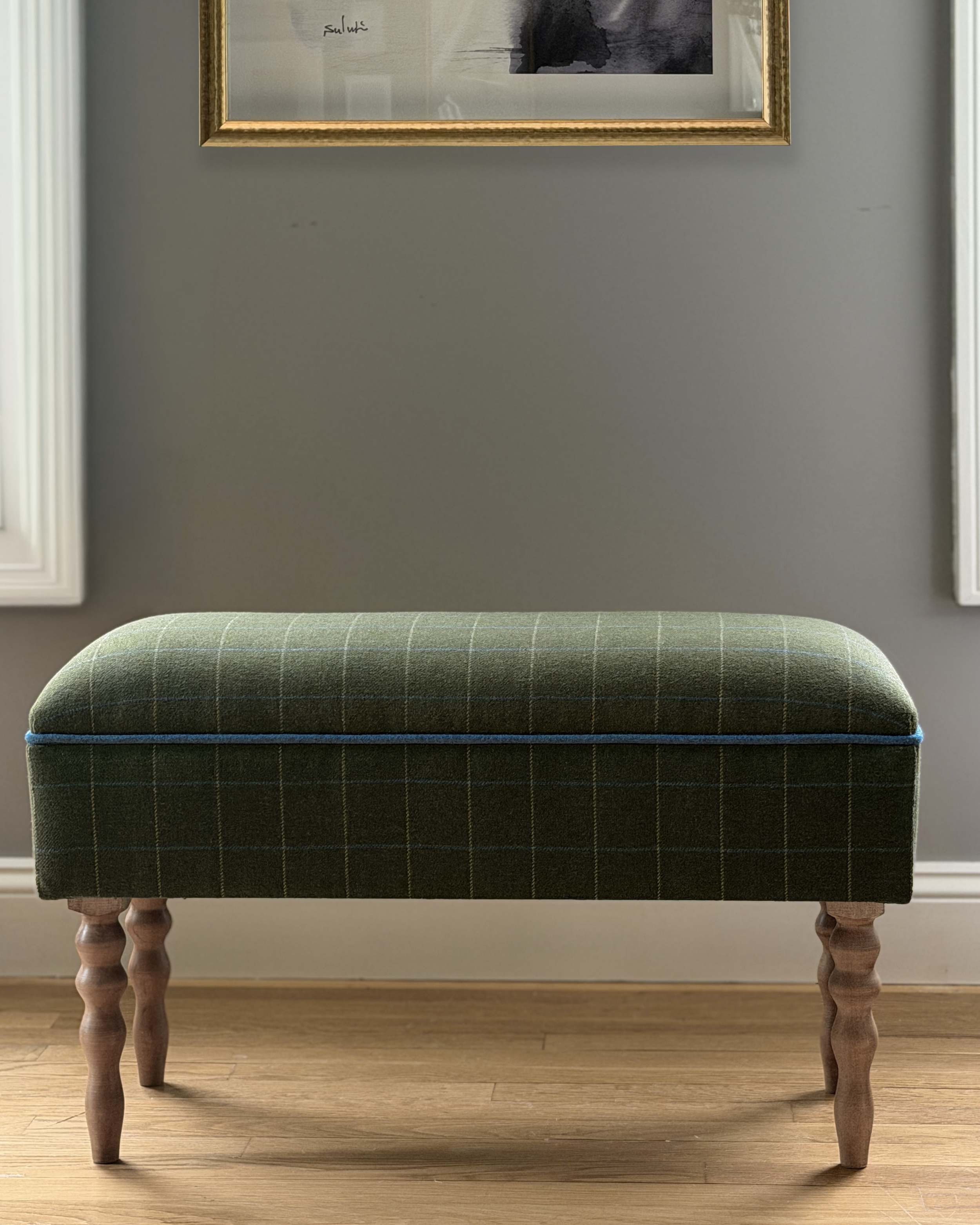 A custom green upholstered ottoman with wooden turned legs in a room with light hardwood flooring, positioned in front of a gray wall. A framed black-and-white abstract artwork hangs on the wall above the ottoman.