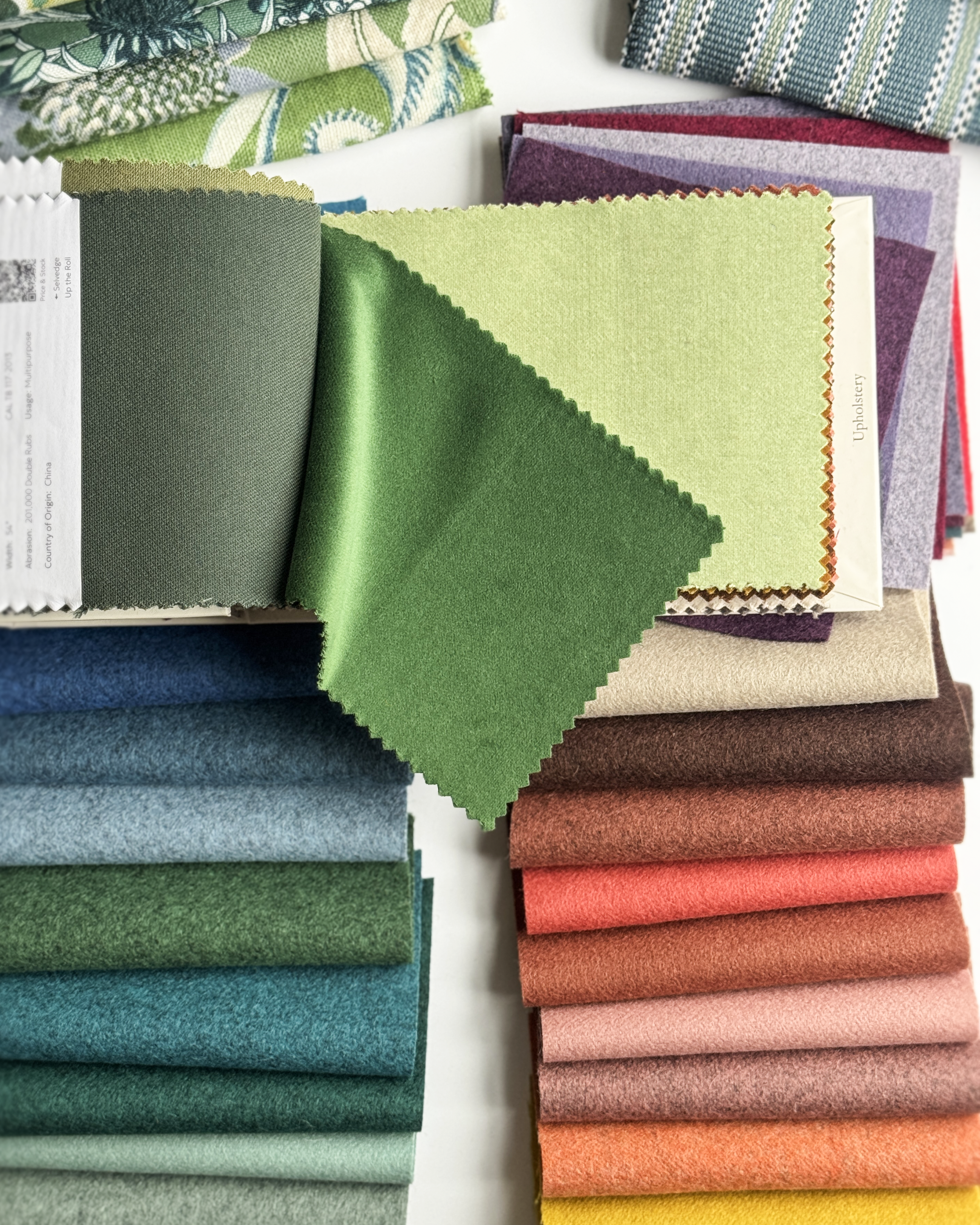 A collection of fabric swatches in various colors, including greens, blues, purples, reds, pinks, and yellows, arranged in two stacks on a white surface.